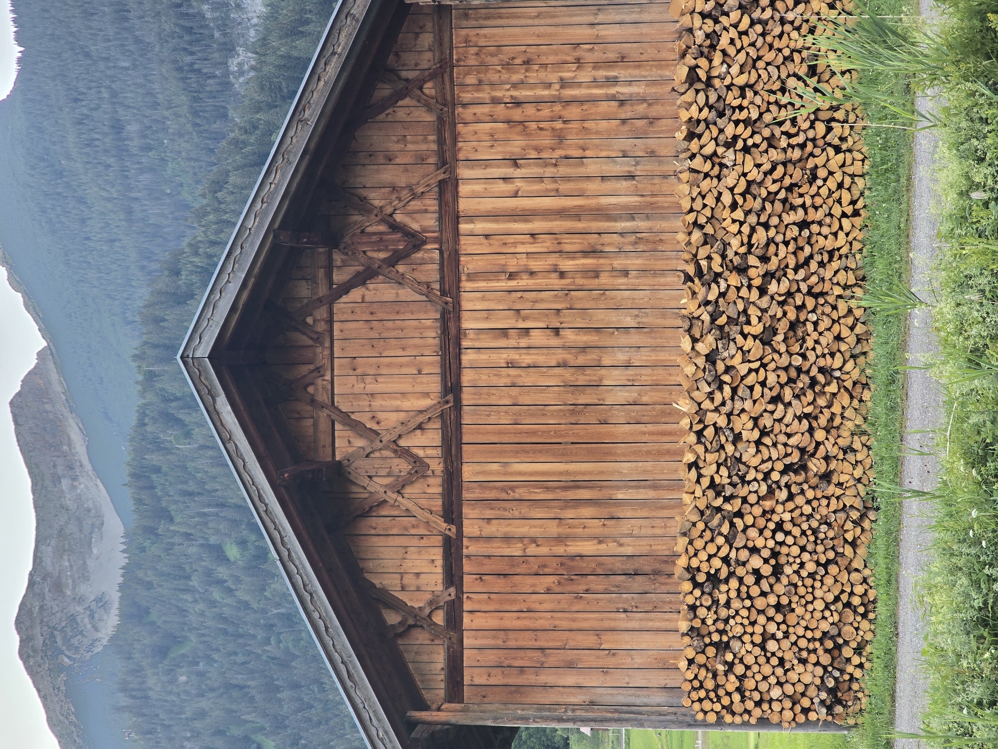 A full-frame, eye-level, vertical shot depicts the wooden facade of a barn. The barn is constructed of vertical wooden planks, with a triangular section at the top featuring decorative wooden crossbeams. A large pile of neatly stacked firewood occupies the lower portion of the barn's wall. Behind the barn, a dense forest of green trees covers a mountain slope, and the peak of a gray, rocky mountain is visible at the top of the image. In the foreground, lush green grass and a gravel path are present. The scene is set in Lermoos, Austria.
