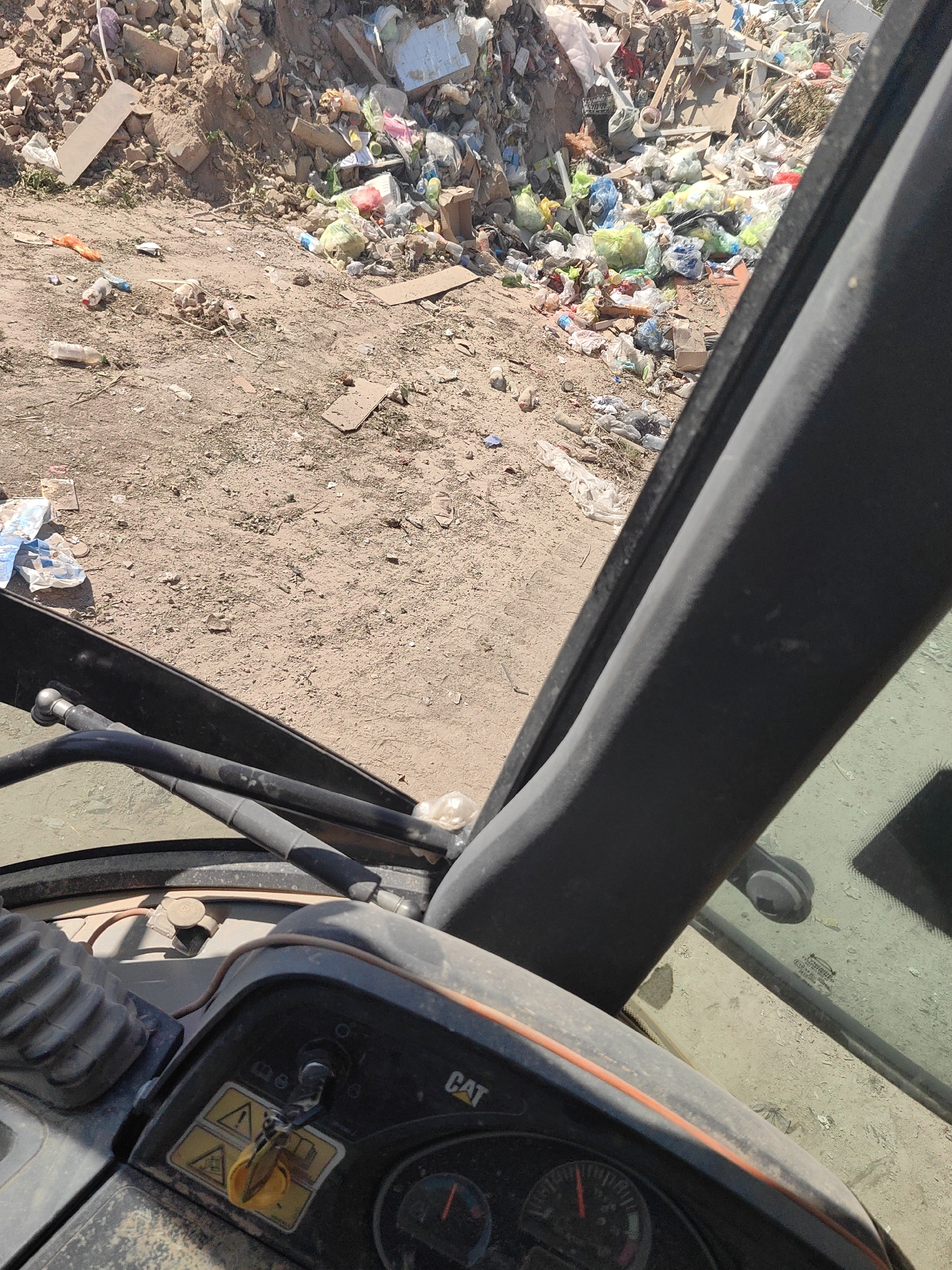 The image was captured inside a CAT excavator cabin, looking out at a landfill in Shymkent, Kazakhstan. The landfill is a large pile of assorted trash, including plastic bags, bottles, cardboard, and construction debris. The ground around the landfill is dry and dusty. The excavator cabin is equipped with a steering wheel, levers, and a control panel with various gauges and warning symbols. The overall scene depicts a working environment within a waste management facility.