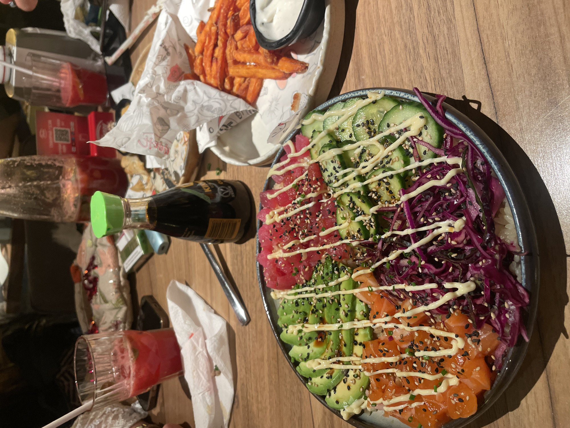 A high-angle medium shot shows a table arrangement of food and drinks, seemingly in a restaurant. A large, deep bowl sits in the center-right of the image, containing a colorful poke bowl with ingredients like tuna, avocado, cucumber, salmon, and red cabbage, drizzled with sauce and sprinkled with sesame seeds. Next to it on the left, there's a plate of sweet potato fries with a small bowl of white sauce. To the left of the fries, there is a bottle of Kikkoman soy sauce and a glass of red liquid. The table surface is light brown wood. The location is likely Sofia, Bulgaria.