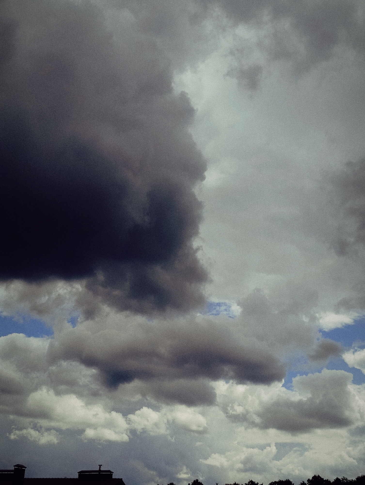 The image shows a sky filled with dramatic, dark storm clouds. The clouds are a mixture of deep grays and blacks, with lighter, wispy clouds interspersed throughout. A sliver of bright blue sky is visible in the upper right corner, suggesting a break in the storm. In the foreground, silhouetted against the sky, are the tops of buildings and trees. The roofs have chimneys, indicating an urban or suburban setting. The overall impression is one of an impending storm.