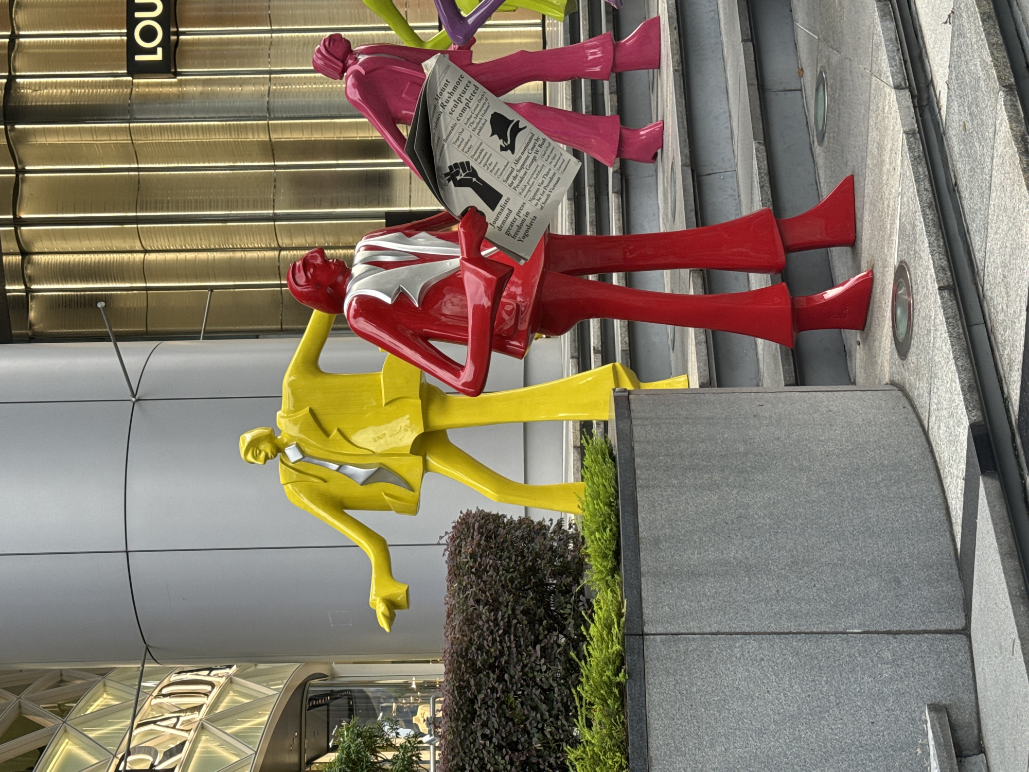 This image depicts a public art installation in front of the Louis Vuitton Island Maison and Prada Epicenter in Singapore. The installation features several brightly colored, stylized human figures made of polished material. In the foreground, a figure dressed in red stands holding a newspaper, its pages detailing news articles and headlines such as "Mount Rushmore sculptures completed" and "Journalists demand greater press freedom in Yugoslavia." Behind the red figure, a yellow figure stands with its arms outstretched, and further back, a pink figure is visible. The figures are positioned on tiered steps made of stone, with lush green bushes and shrubbery bordering the lower levels. The architectural background includes modern buildings with sleek facades and illuminated panels, indicative of a high-end retail district. The lighting suggests it is daytime, with natural light illuminating the scene.