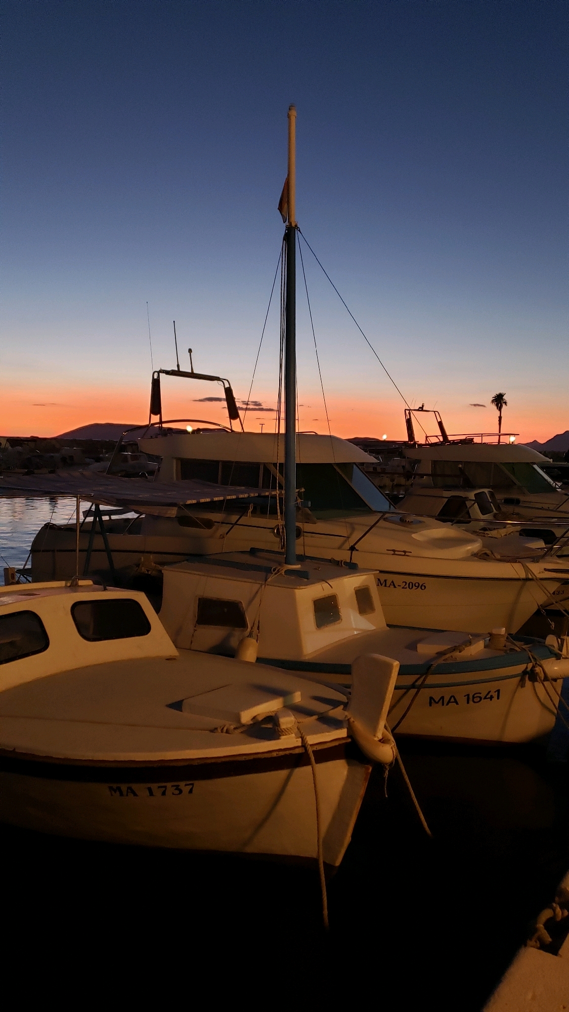 The image depicts a harbor at dusk, with a gradient of blue to orange in the sky. Several boats are moored, their structures silhouetted against the fading light. In the foreground, two white boats are prominently visible. The boat on the left has the registration number "MA 1737" on its hull. To its right, another white boat, partially obscured, shows the registration number "MA 1641". Behind these, larger boats with cabins and masts are docked. A single palm tree is visible on the distant shoreline, which is marked by low hills or mountains. The water is calm and reflects the sky. The scene suggests a peaceful evening in a coastal town. This scene is consistent with the harbor area of Općina Tučepi, Croatia, known for its picturesque Adriatic coastline and marina.