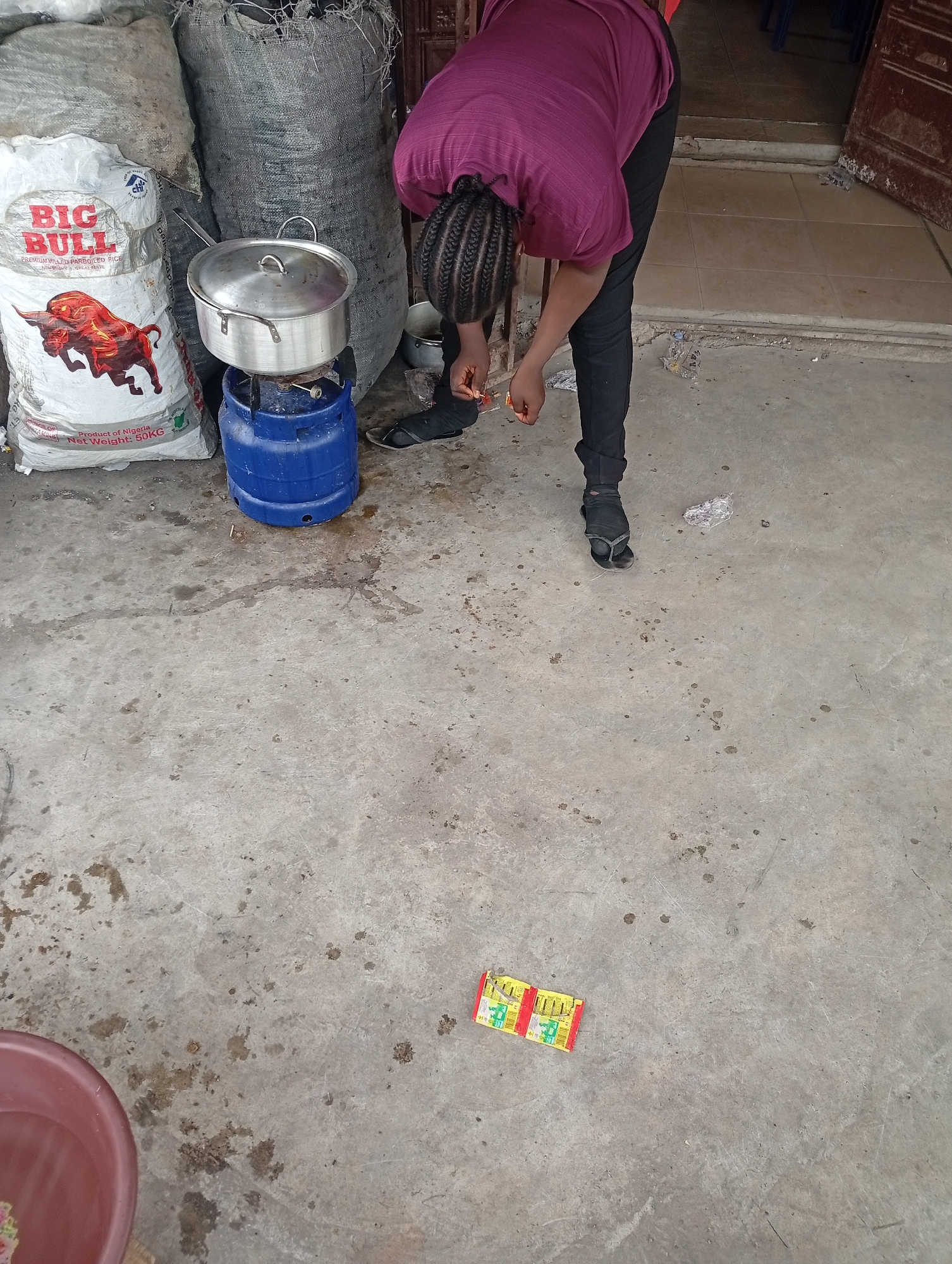 This image captures a person in an outdoor or semi-outdoor setting, engaged in a cooking-related activity. A person, with braided hair, wearing a maroon top and black trousers, is bent over, their hands near the ground, possibly opening small yellow packets. They are standing near a cooking setup consisting of a blue gas cylinder with a silver pot on top.
To the left of the person, a large white sack of "BIG BULL PREMIUM MILLED PARBOILED RICE" is visible, featuring a red bull logo. The sack states "Product of Nigeria" and "Net Weight: 50KG." Two other large, dark grey/black sacks lean against a wall behind the rice sack. The ground is a stained concrete floor, and a red plastic basin is partially visible in the lower-left corner. Two identical yellow packets are also on the floor in the foreground. The background shows a doorway leading to an indoor space with tiled flooring. The scene appears to take place during the day under diffused light. This setup suggests a common domestic or small business cooking area in a Nigerian context, aligning with the Adankolo, Nigeria location.