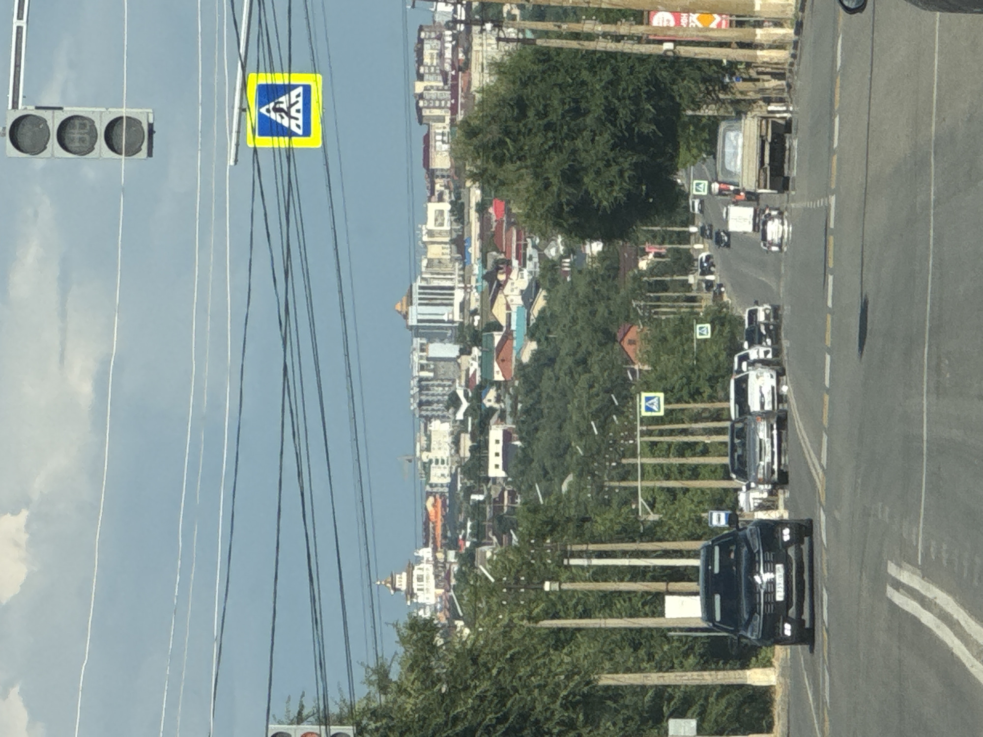 The image shows a street scene in a town or city, likely in Russia, as indicated by the "Дорожный" context. The photograph is taken from a slightly elevated perspective, looking down a road lined with trees and buildings. The road appears to be a two-lane road with a white dashed line dividing the lanes. Several vehicles are visible on the road, including cars and a truck, suggesting that this is a moderately busy street. One black SUV in the foreground has a license plate that reads "T239CA 777". There are also pedestrian crossing signs visible, indicating that this is a safe area for pedestrians to cross.
The buildings lining the street appear to be a mix of residential and commercial structures, with various architectural styles and colors. Some buildings have multi-story designs, while others are smaller, more traditional homes. Lush green trees line the sides of the road and are interspersed with the buildings, adding a natural element to the urban landscape.
In the upper left corner, a traffic light with three circular lights is visible. Next to it, a yellow square sign with a blue triangle and a white symbol is mounted on a pole. Overhead, numerous electrical wires crisscross the sky, which is partially obscured by clouds, suggesting it might be a partly cloudy day.
The overall impression is a typical day in a Russian town or city, with everyday activities like driving and commuting taking place. The lighting suggests it's daytime, possibly late morning or early afternoon, given the bright sunlight and clear shadows. There are no discernible emotions expressed, as the scene is focused on the environment and the objects within it. The image is captured in a vertical orientation.