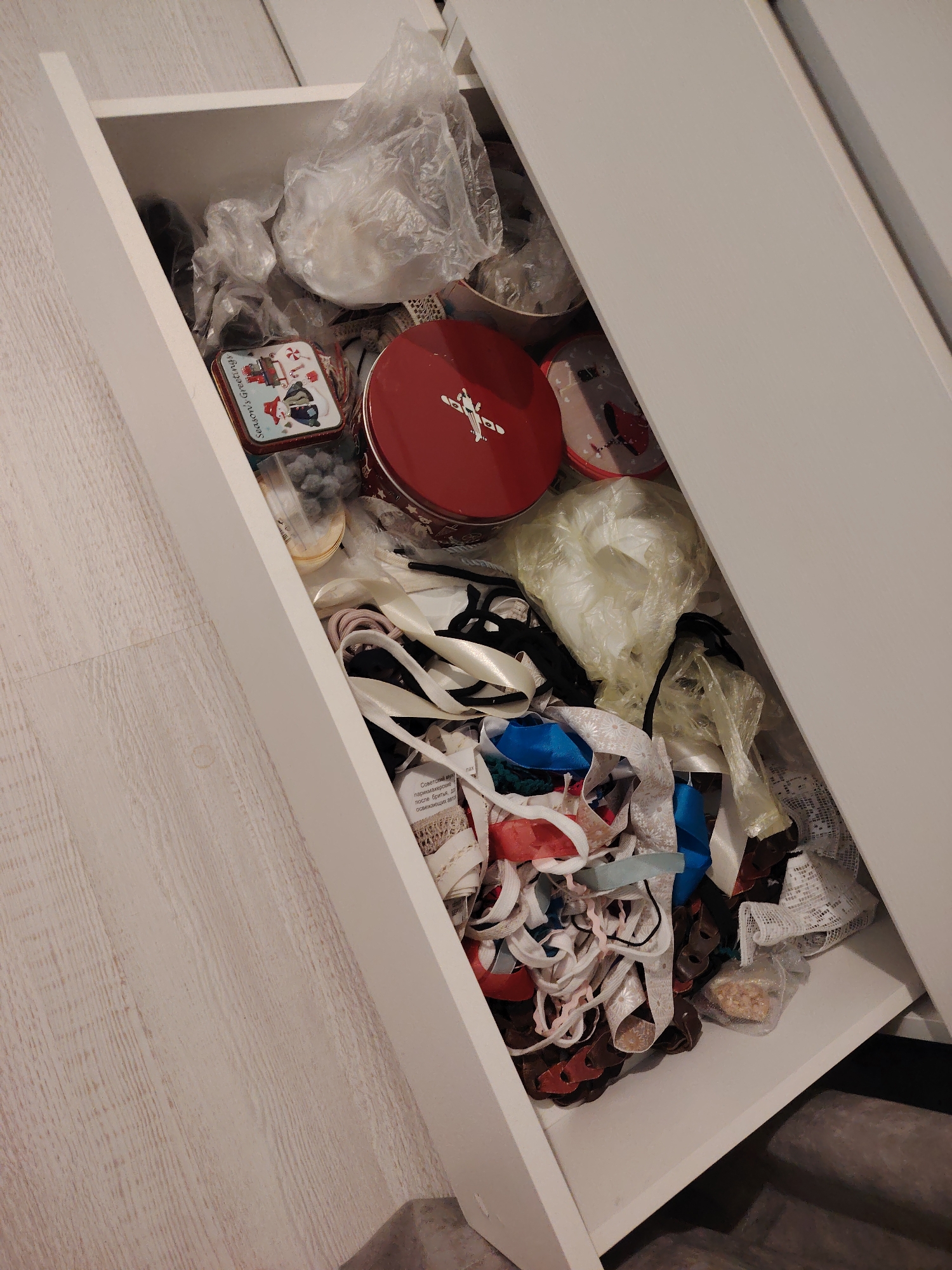 An overhead view captures a partially open white drawer filled with various items, suggesting a collection of craft supplies or miscellaneous household storage. The drawer contents appear somewhat disorganized.
Dominating the upper section of the drawer are several clear plastic bags, some containing white or light-colored textiles or stuffing. Towards the top left, a small, rectangular metal tin is visible, decorated with holiday figures like snowmen and houses, and bears the text "Season's Greetings." Adjacent to it is a larger, round red metal tin with a white stylized design, possibly a reindeer or other holiday motif. Below these tins, a clear container holds small grey pom-poms or similar craft embellishments.
The lower half of the drawer is densely packed with an assortment of ribbons, sashes, and lace trimmings of various widths and materials. Colors include white, cream, black, blue, red, and brown. Some pink-toned hair ties or scrunchies are also present among the ribbons. A label attached to some white lace or ribbon has partially visible Cyrillic text, including "Советский" (Soviet) and "парикмахерские" (hairdressing), suggesting a product description. Another small plastic bag containing light-colored granules or small items is visible at the very bottom right of the drawer.
The drawer itself is white, and the surrounding floor is a light wood-grain laminate or planking. A darker, possibly carpeted, surface or steps is visible below the drawer opening on the right side. The lighting suggests an indoor setting, likely under ambient room light. No specific activity is depicted, only the state of the stored items.