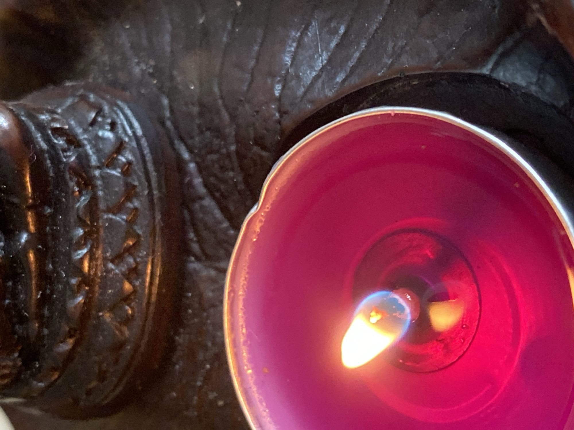 A top-down close-up shot shows a burning pink candle in a silver holder next to a carved dark brown object. The candle flame is visible in the center of the pink wax. The carved object appears to be resting on a dark, textured surface. The image does not provide any evidence that it was taken in Baan Kai, Thailand.