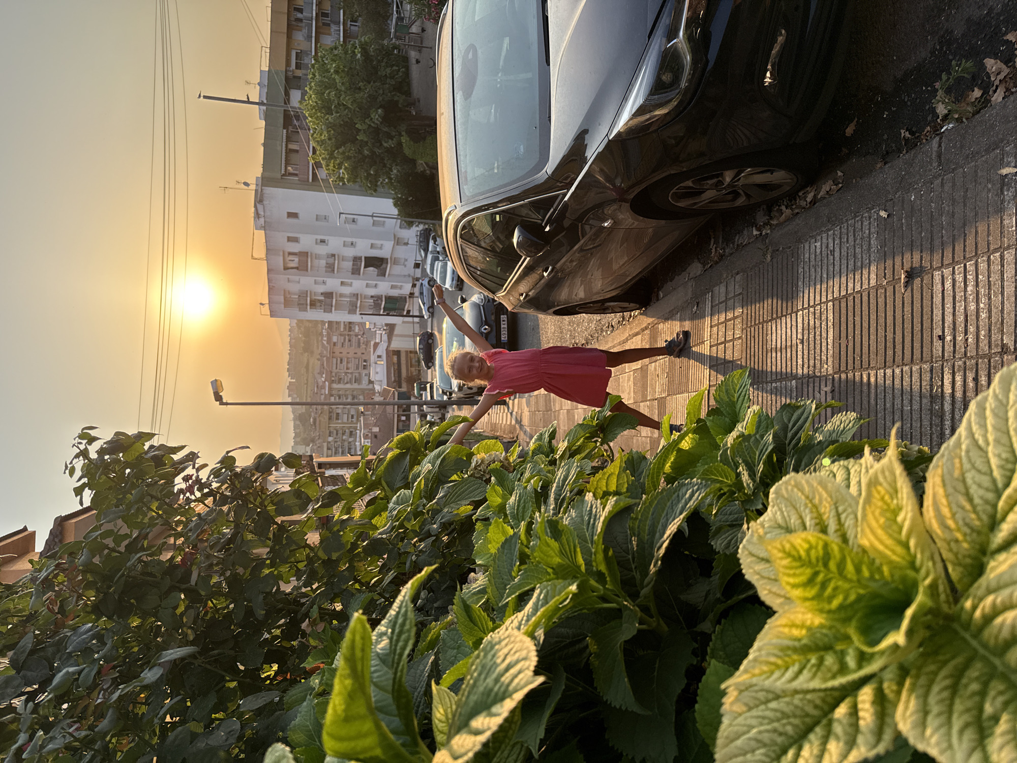 A young girl with blonde hair, wearing a pink dress and sneakers, stands on a sidewalk in Blanes, Spain, with her arms outstretched. The sun is setting, casting a warm golden glow over the scene. To her right, a dark-colored car is parked on the street. In the foreground, lush green plants are visible. Behind the girl, residential buildings and trees line the street. The overall impression is one of a peaceful evening in a Spanish town.