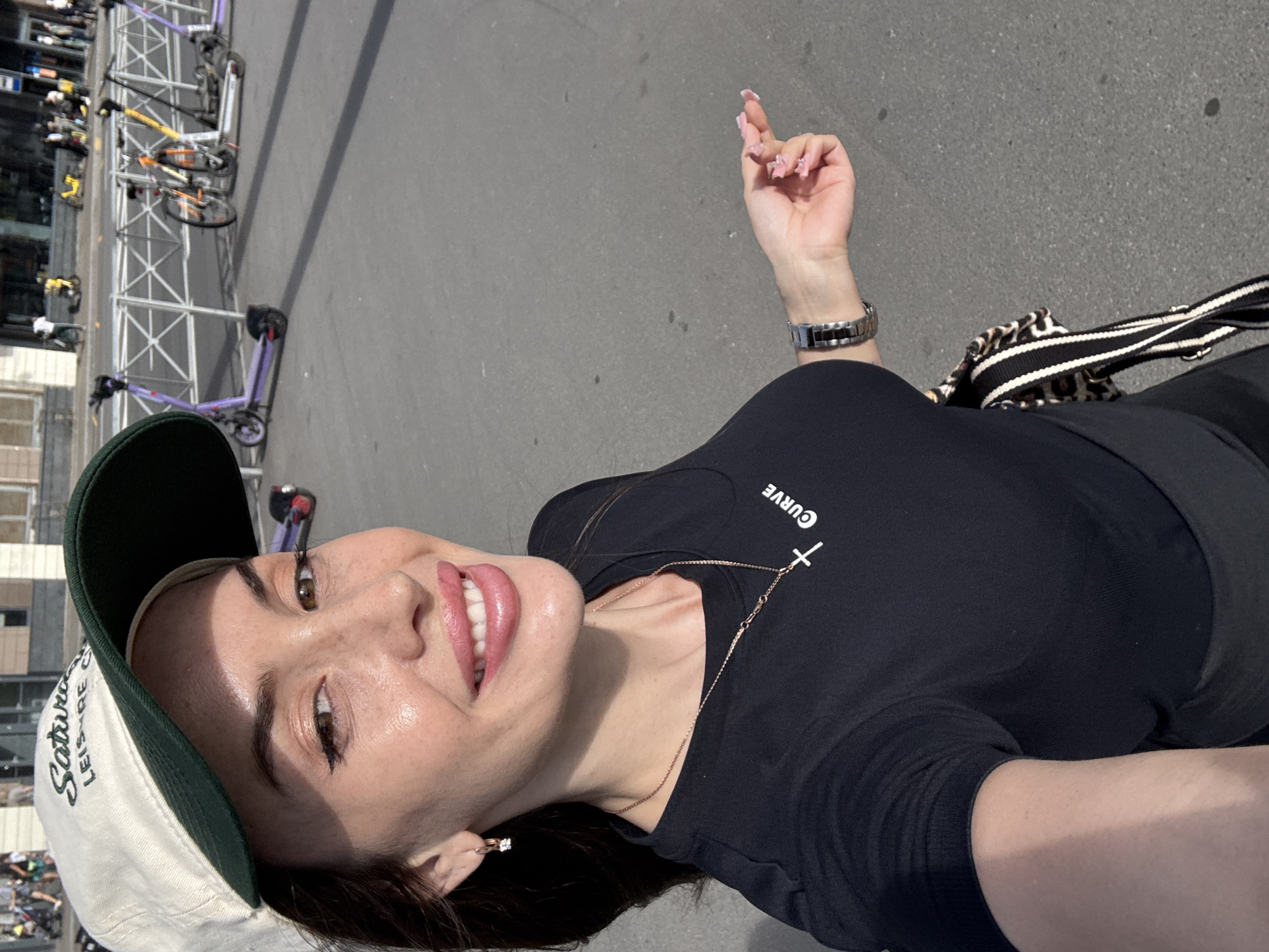 A woman is taking a selfie outdoors, with her head tilted back and a bright smile on her face. She is wearing a black t-shirt with the word "CURVE" and a cross symbol printed on it. A gold chain with a cross pendant rests on her collarbone. She has on a baseball cap, green on the brim and white on the crown, with some lettering visible. She is also wearing a silver watch on her left wrist. In the background, a row of scooters and bicycles are parked on a paved surface, suggesting an urban setting. The sunlight suggests it is daytime, and the shadows indicate it is likely a sunny day. The woman's expression conveys happiness and a sense of enjoyment.