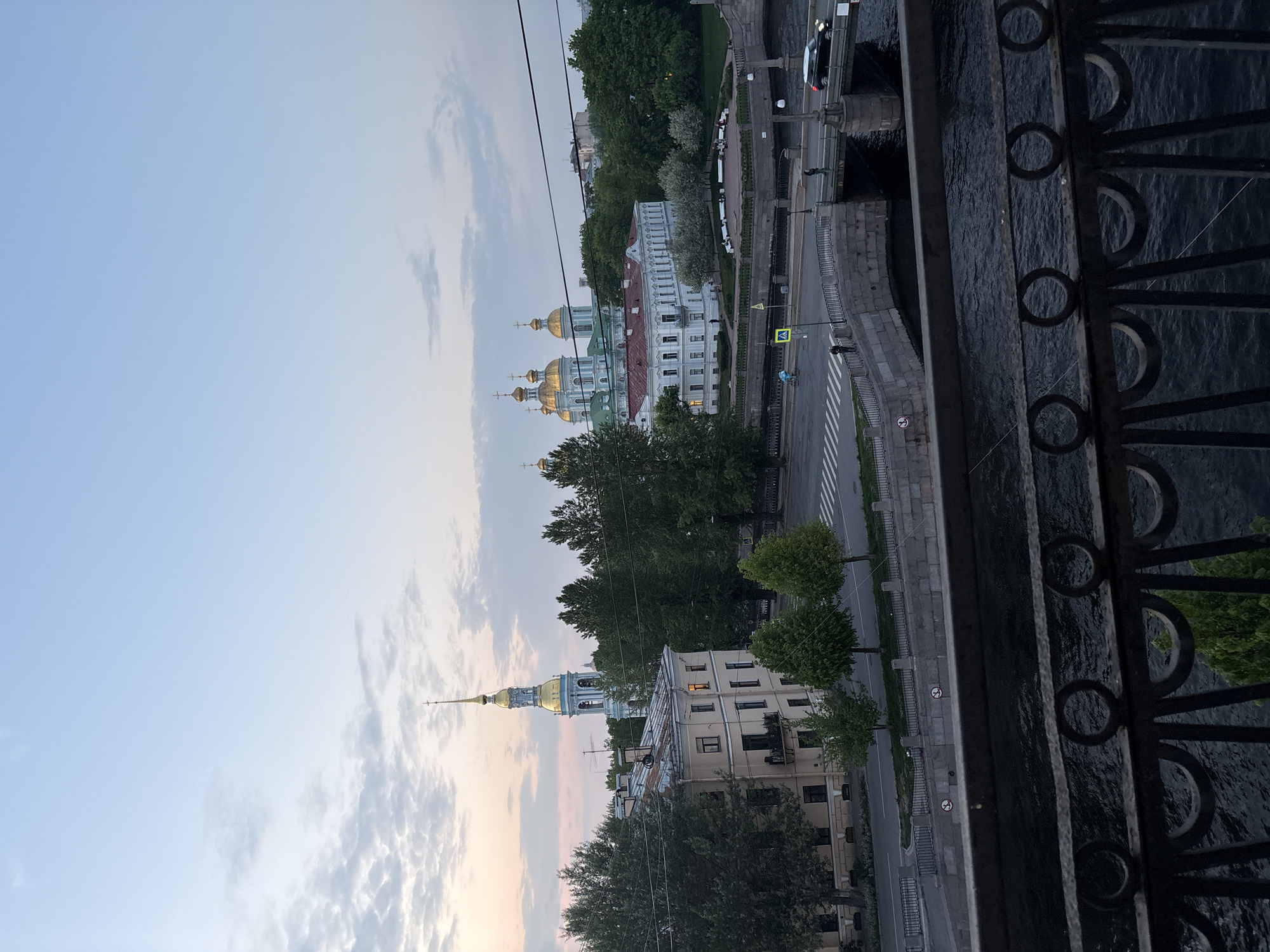 The image depicts a view from a bridge in Saint Petersburg, Russia. The sky is a pale blue with soft clouds. In the distance, there are two prominent landmarks: the Smolny Cathedral and the Saint Sampson's Cathedral, both identifiable by their gold domes. A tree line separates the buildings from the roadway. A crosswalk is visible on the road, and some cars and pedestrians are also visible. The foreground features a black wrought-iron fence with circular elements. The river flows to the right of the frame.