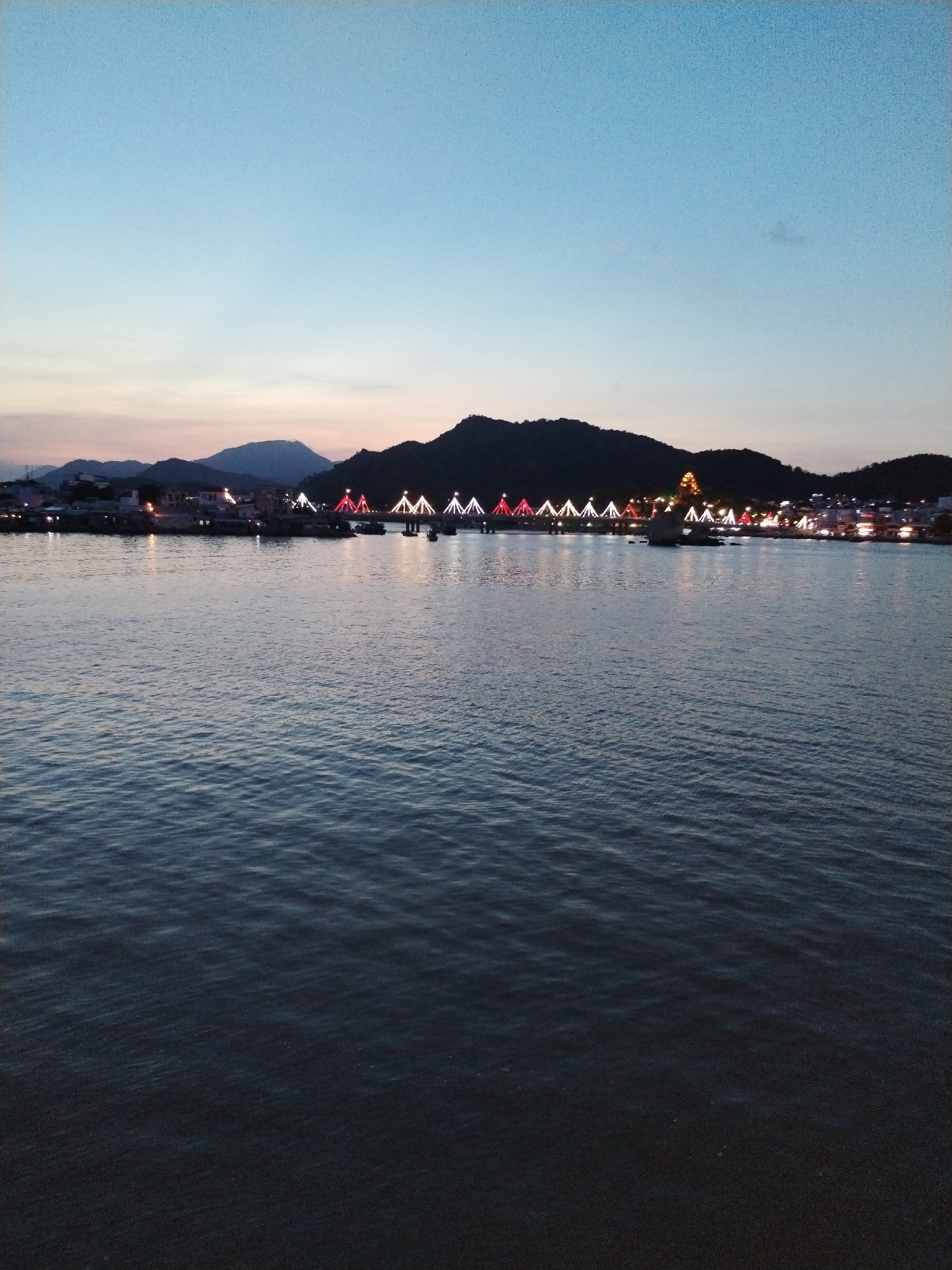 The image captures a waterfront view in Nha Trang Ward, Vietnam, at dusk. The foreground is dominated by the calm, dark water of the bay, reflecting faint light. In the midground, a series of buildings and structures line the opposite shore. A bridge spans the water, illuminated with alternating red and white lights, creating a visually striking feature. Behind the developed shoreline, a dark mountain range rises against a fading blue sky. The overall scene presents a tranquil yet urban waterfront environment.