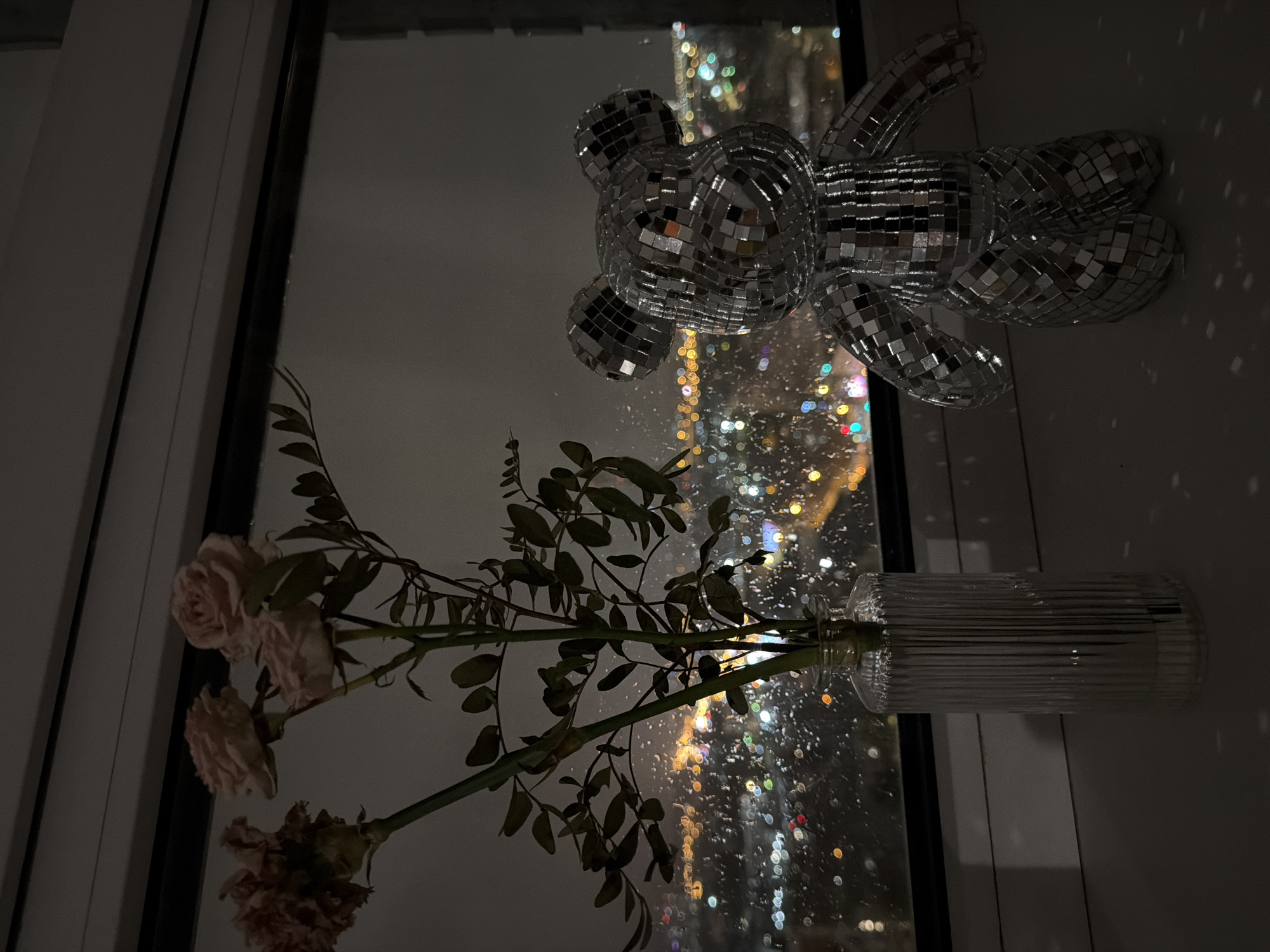 The image shows a window with a cityscape visible through the glass. A disco bear and a vase with flowers are positioned near the window. The cityscape is illuminated with many lights, suggesting it is night. The disco bear reflects the cityscape lights. The flowers in the vase are pale pink and some appear to be wilting.
