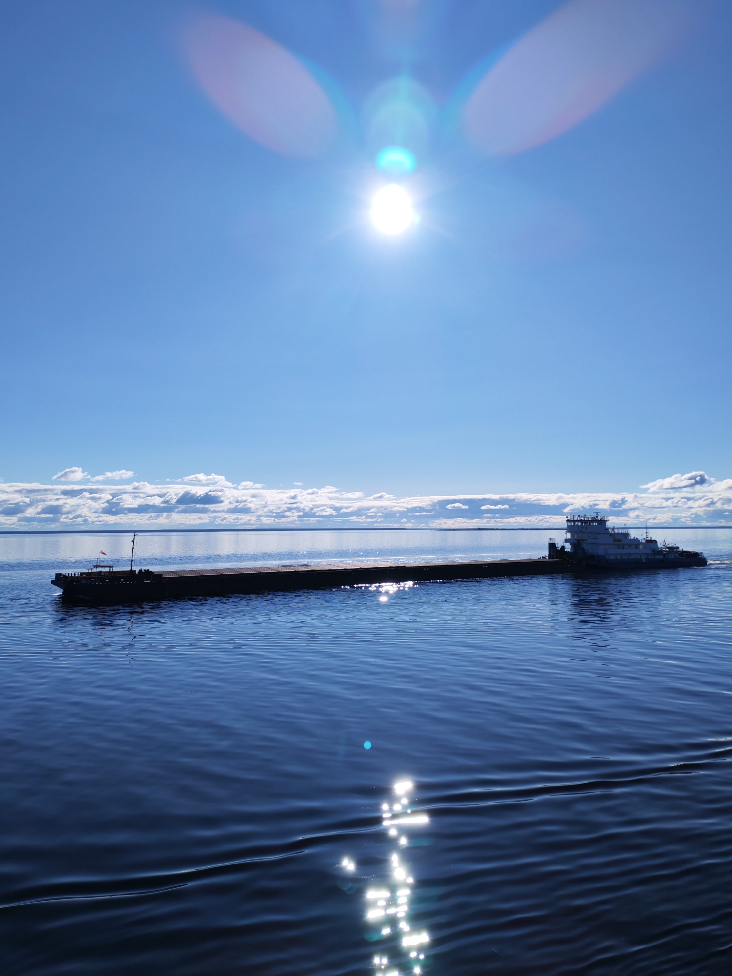 A tugboat and a long barge are traveling on a body of water under a bright, sunny sky. The water is reflecting the sunlight. There is a low-lying land mass or shoreline in the distance with clouds above it. The location is not specified in the image.
