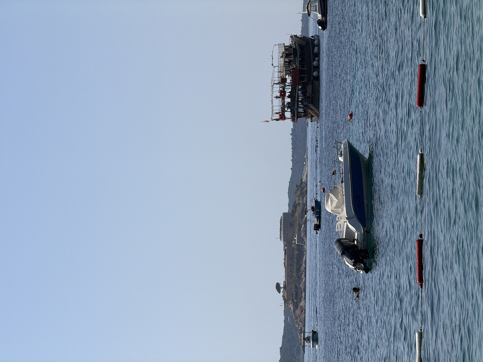 In the city of Luštica, Montenegro, a body of calm, blue water is dotted with various vessels under a clear, bright sky. In the foreground, a white motorboat with a blue stripe and a dark outboard motor is moored. A person with a snorkel is visible in the water nearby. Further out in the water, a large, dark, multi-decked tourist boat with many passengers is cruising. In the mid-ground, several smaller motorboats are scattered across the water. In the background, a fortified island complex with a prominent round tower and a lone tree on its highest point sits on a rocky outcrop. The shoreline behind the island is lined with green hills. A rope with alternating white and red buoys stretches across the very foreground of the image, delineating a swimming area.