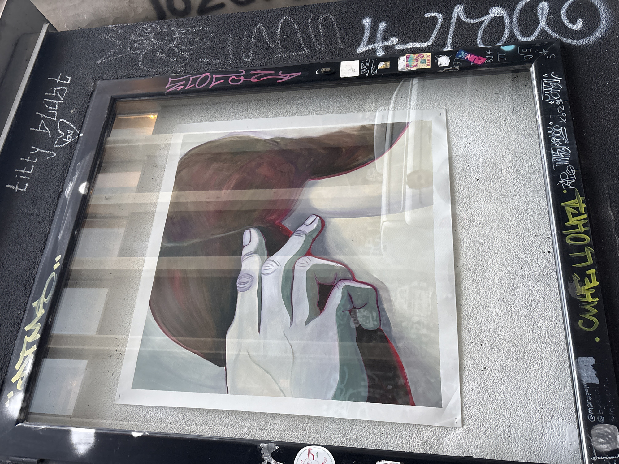 A framed poster is mounted on a textured, light-gray wall. The poster depicts a painted image of a hand touching a breast. The frame is black and covered in graffiti tags and stickers. "titty blast" is written on the left side of the frame with a heart drawn between the words. The top of the frame has various tags and stickers, as well as white graffiti. The right side of the frame also has graffiti tags, written in both green and white. The surface above the frame also has more graffiti. The location is likely Belgrade, Serbia based on some of the graffiti.