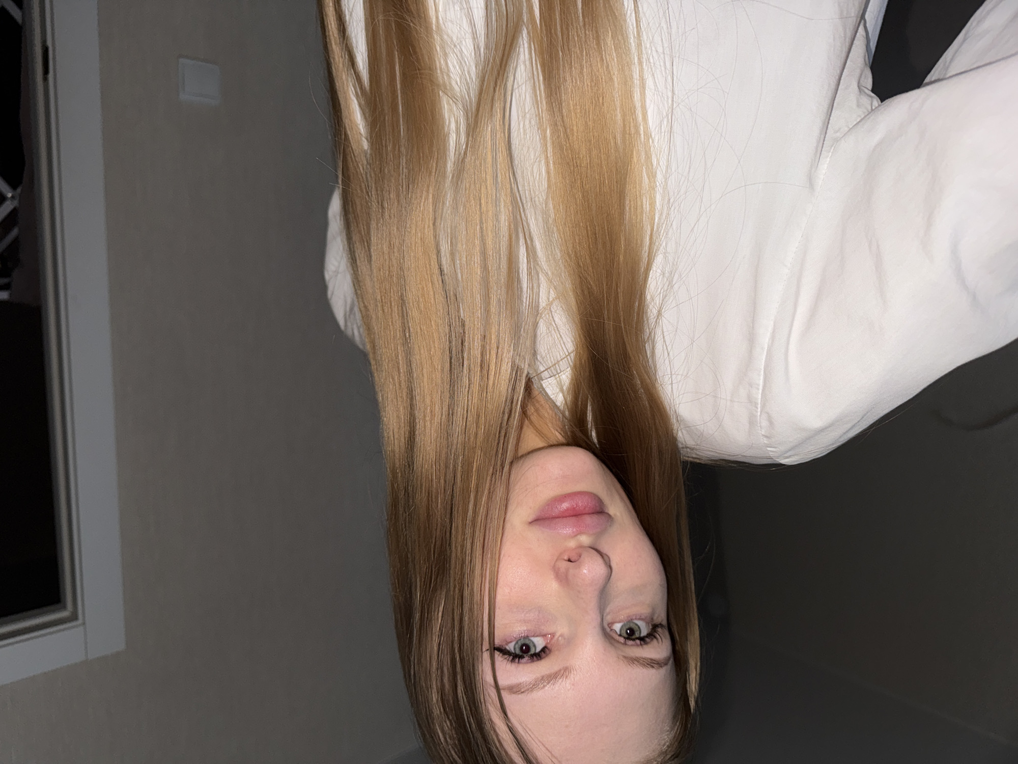 The image shows a young woman with long, blonde hair, wearing a white shirt. She is looking directly at the camera with her head tilted slightly. The background is a plain, light-colored wall. There is a doorway visible to the right of the woman. The lighting is dim, suggesting an indoor setting, possibly at night. The image is a selfie, taken from a slightly low angle. The woman has fair skin, light blue eyes, and full lips. She is wearing subtle makeup, including eyeliner and mascara. The overall impression is a casual, personal photograph. There are no specific clues in the image that would allow for its location to be identified as the city of Perm, Russia.