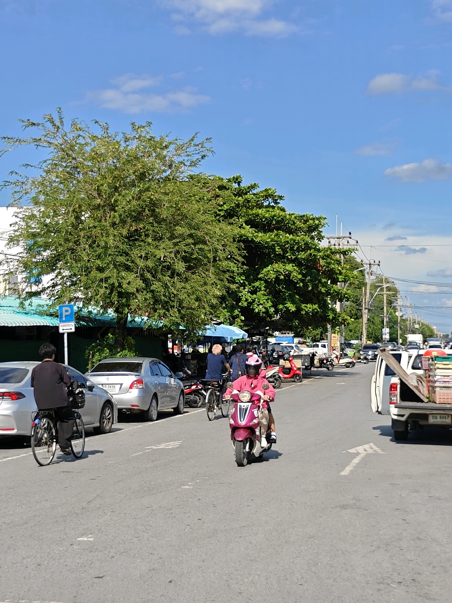 The image depicts a street scene in Phra Nakhon Si Ayutthaya City Municipality, Thailand, under a bright blue sky with scattered clouds. The foreground is dominated by a paved road with a white arrow indicating a turning lane. A white pickup truck with cargo in its bed is parked on the right side of the road. In the middle ground, several people are present. A person on a pink scooter, wearing a pink helmet, is riding down the road. To the left, another person is riding a bicycle. Behind them, various parked motorcycles and cars are visible, along with some street vendors' stalls. A silver sedan is parked on the left, and a silver car with a Thai license plate is in front of it. Further back, more vehicles and buildings line the street, including a building with a green roof and a blue parking sign. Large trees with dense green foliage are situated along the left side of the road, providing shade. The overall scene suggests a typical day on a moderately busy street in a Thai urban environment.