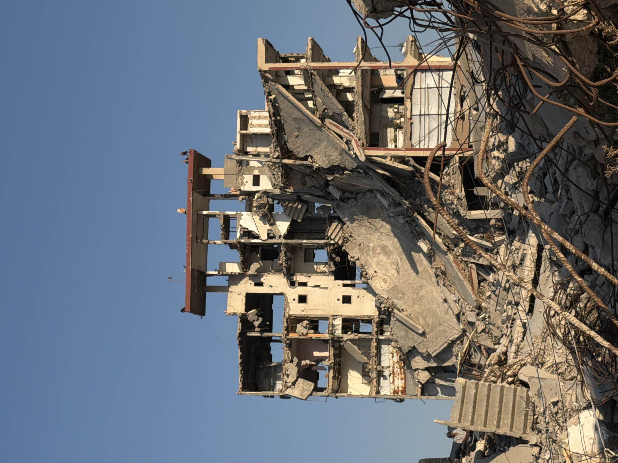 The image shows the heavily damaged remains of a multi-story building in Jabalia, Palestinian Territories, under a clear blue sky. The structure appears to be in a state of ruin, with several floors collapsed and concrete debris scattered around the base. Exposed rebar, twisted and rusted, is visible throughout the wreckage, indicating significant structural failure.
In the foreground, a pile of rubble includes large chunks of concrete and twisted metal. A partially intact concrete staircase ascends from the debris on the left side of the image. A person, possibly a man wearing a light-colored shirt, is sitting on this staircase, looking towards the right.
The building itself is severely damaged, with walls missing and floors exposed. Windows are either broken or absent, and inner structures are visible through the gaping holes. On the uppermost surviving section of the building, a red-tiled roof or canopy is partially visible, with a few birds perched on its edge. Another person, a woman with light-colored hair, is visible standing on this upper level, also looking towards the right. The overall scene suggests destruction and the aftermath of an event that has left the building in a derelict state.
