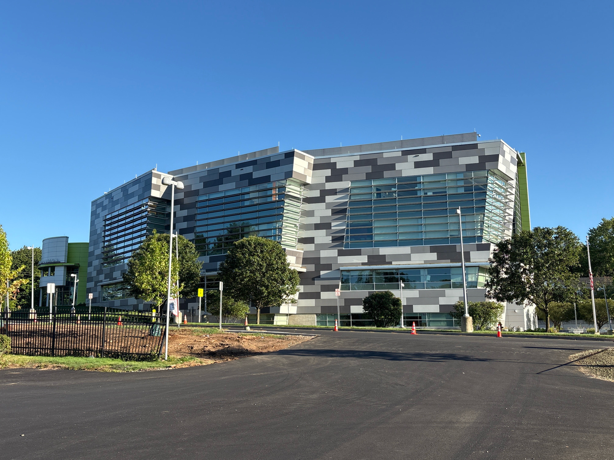 The image shows a modern building with a facade composed of alternating gray and white rectangular panels, interspersed with large glass windows. The building has a distinctive angular design with a prominent green section on the left side. Several trees are visible around the building, and the sky is a clear blue. In the foreground, there is an asphalt surface, likely a road or parking lot, with a black metal fence and some landscaping in progress. There are orange traffic cones placed along the edge of the asphalt and in front of the fence. A few signs and light poles are also present. The overall scene depicts an exterior view of the building, possibly on a sunny day. The building is located at the Connecticut Department of Transportation (CTDOT) District 1 Maintenance Facility in East Hartford, Connecticut, United States of America.
