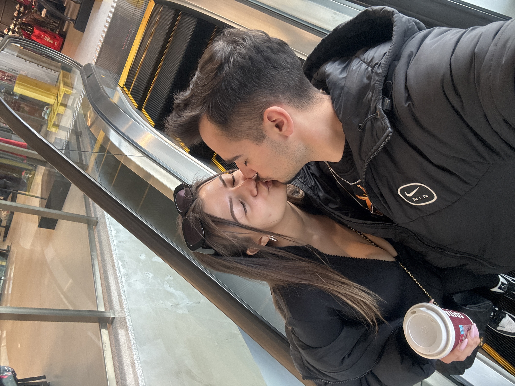 The image captures a tender moment between a man and a woman kissing on an escalator, likely within a shopping mall or large public building in Sofia, Bulgaria. The photograph is taken at a diagonal angle, emphasizing the intimate interaction.
The man, with dark, styled hair and light stubble, wears a black puffer jacket featuring a white Nike swoosh logo and the word "AIR" on the sleeve. A silver chain is visible around his neck. He is leaning down to kiss the woman, whose eyes are closed in the embrace. She has long brown hair, with dark sunglasses pushed up onto her head. She wears a dark top and a dark jacket, and her nails are painted light pink. In her right hand, she holds a white takeaway cup with the faint "Costa" logo visible. Part of her black and white sneakers, possibly Air Jordans, can be seen near the escalator steps.
The setting is an indoor environment, characterized by the modern escalator with metal handrails, transparent glass panels, and black steps highlighted with yellow edges. Reflections of the surroundings are visible in the glass. In the background, what appears to be shop displays or storage is visible, including a red bag with "Wilson" printed on it. The overall lighting suggests a brightly lit interior, typical of daytime mall operations.