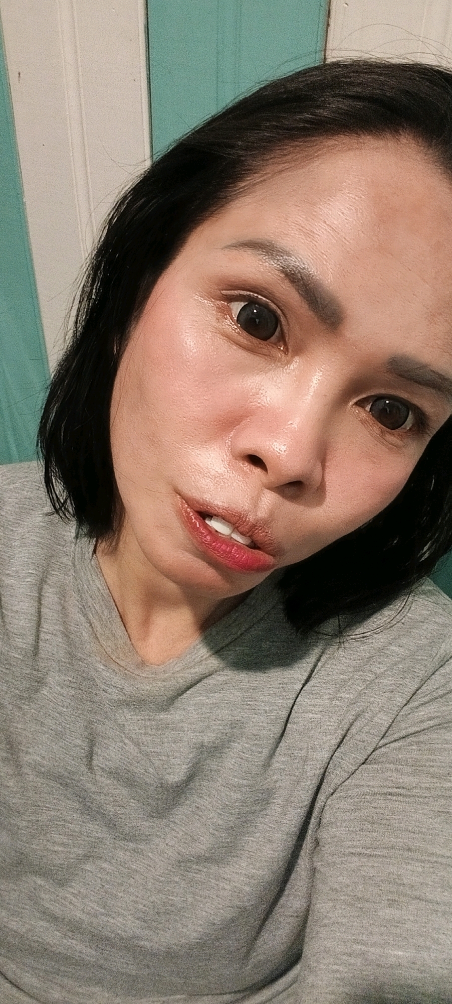 A close-up, over-the-shoulder selfie shows a woman from the chest up, facing the camera. She has black hair, dark eyes, and is wearing a grey t-shirt. Her skin appears shiny, possibly from makeup or sweat. She is looking slightly to her right, with her mouth slightly open, revealing her teeth and lips with a glossy red lipstick. The background consists of a teal-colored wall with vertical white panels. The lighting casts shadows on the woman's face and neck. The image was taken indoors.