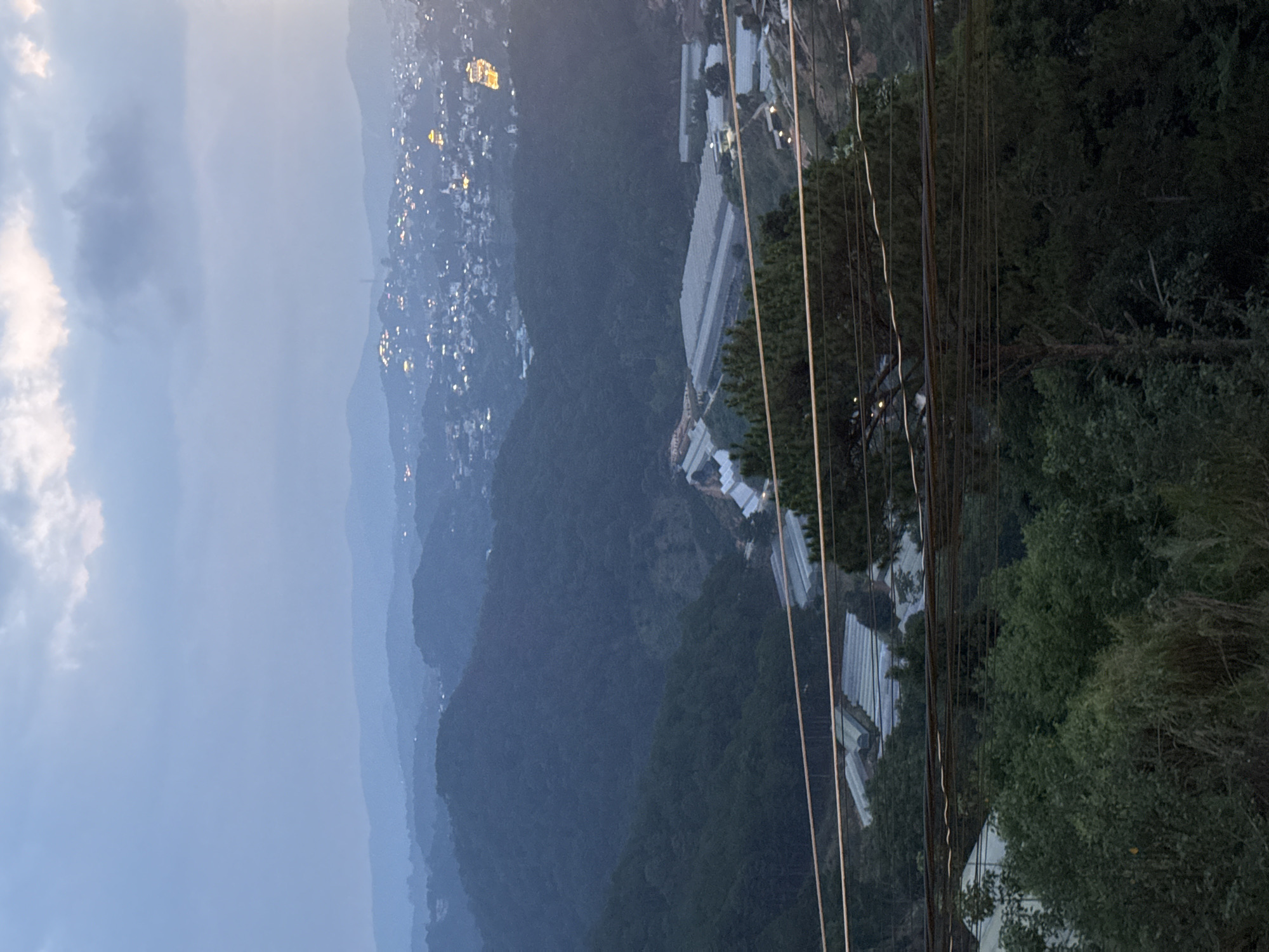 An aerial view shows a cityscape nestled in a hilly terrain at dusk or night. Small, bright lights dot the distant hills, suggesting a residential area. Close by, on the right, structures with white roofs, possibly greenhouses or agricultural buildings, line the slope. Vertical lines, likely power lines or cables, run down the right side of the image. The sky is overcast with clouds visible on the left. Trees and vegetation are visible on the right, adding to the landscape. The location is Xuân Trường in Đà Lạt, Vietnam.