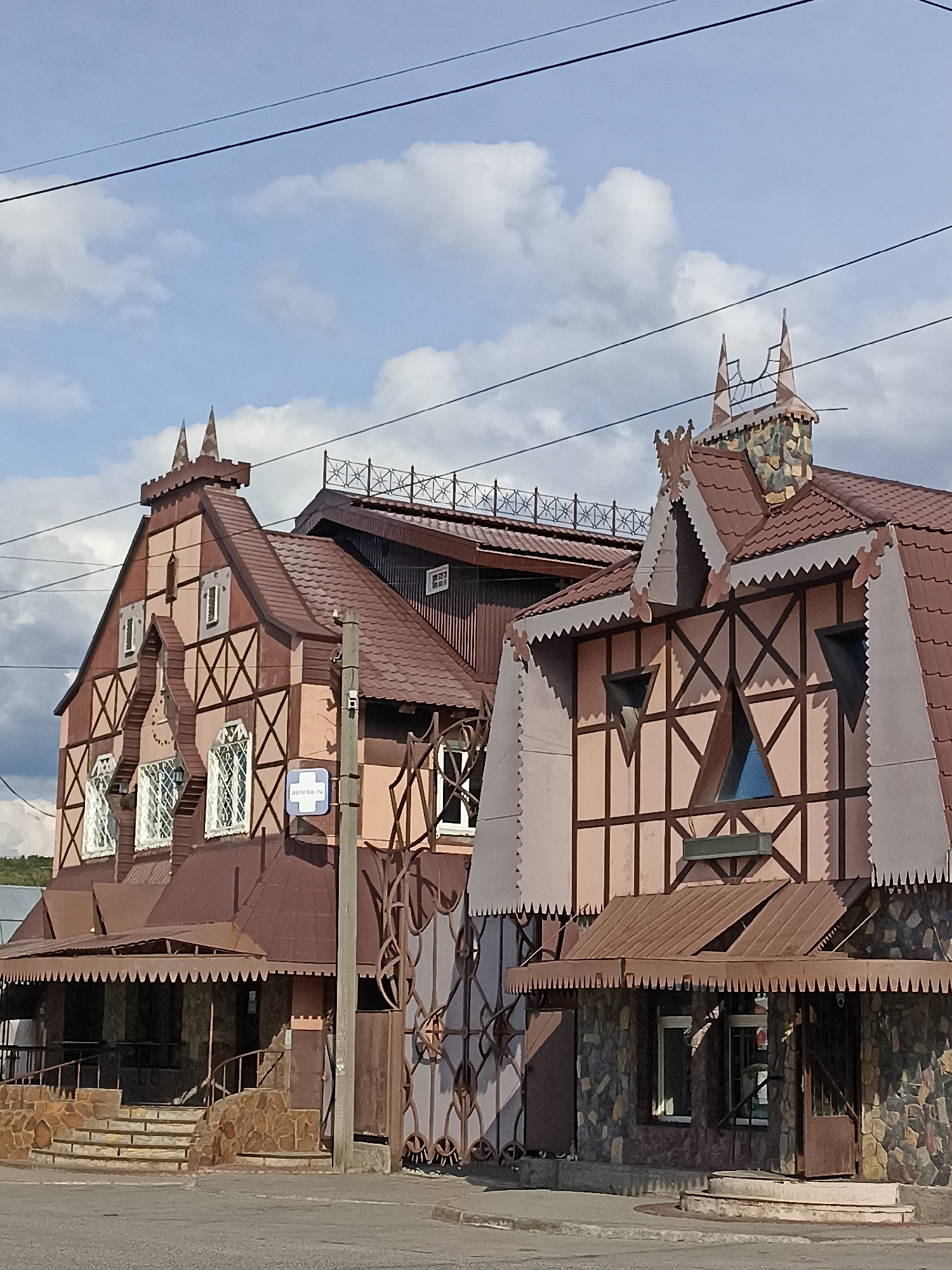 The image shows a street view of two buildings with distinctive architectural styles in Mochische, Russia. The buildings feature a combination of Fachwerk (half-timbered) elements with light pink or beige walls and dark brown decorative beams arranged in diamond patterns. Both structures have steep, brown tiled roofs with decorative gables and finials. The building on the left has multiple windows with white lattice work. The building on the right has triangular windows and a stone-clad lower section. A brown awning with a scalloped edge extends over the entrance to the building on the right. A stone staircase leads up to the entrance of the building on the left. A utility pole with a blue and white sign that says "apteki.ru" is visible in front of the buildings. Decorative metalwork, resembling intertwined branches, is present between the two buildings. Power lines are visible in the sky above. The sky is partly cloudy with blue patches. The ground in front of the buildings appears to be an asphalt road. There are no people visible in the image.
