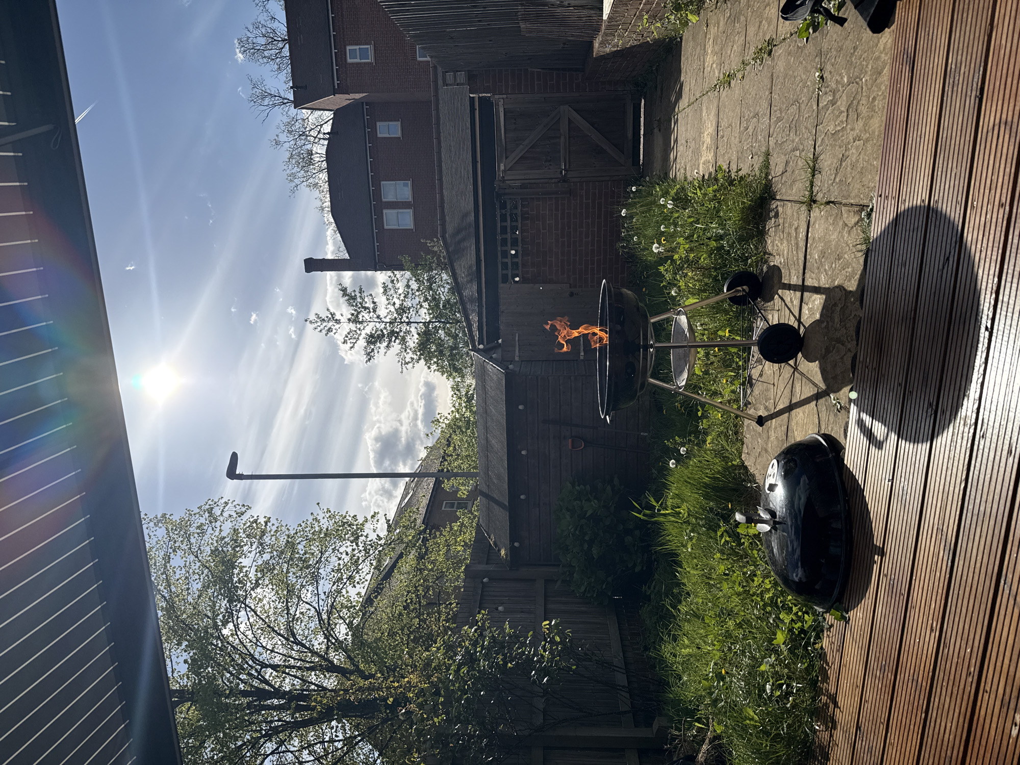 The image shows a garden in London, United Kingdom on a sunny day. A black barbecue grill is burning with flames visible inside. Another black barbecue grill sits nearby on a wooden deck. The garden is bordered by a wooden fence, with a brick building and a shed visible in the background. The ground is a mix of paving stones and grass, with some weeds growing. A street light is visible in the distance.