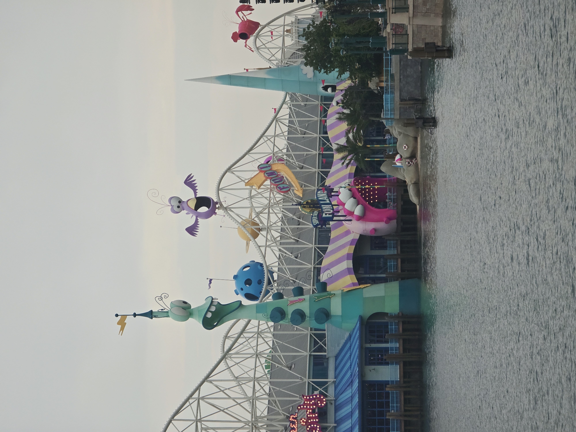 The image captures a waterfront view of an amusement park, likely Universal Studios Beijing, located in the Tongzhou District of China. The sky is overcast, casting a soft light on the scene. In the foreground, a body of calm water reflects the structures and the sky.
Dominating the mid-ground is a roller coaster with a white, lattice-like structure. Scattered across the roller coaster's supports and at the top are colorful, cartoonish figures. One prominent figure on the left is a tall, green, alien-like character with large, staring eyes and an open mouth. To its right, a purple, winged creature with wide eyes is perched. Further to the right, a red, crab-like creature with pincers is visible.
Beneath the roller coaster, an amusement park building with a blue roof and large windows is situated along a wooden pier. A striped awning, featuring purple, yellow, and white bands, extends from the building. To the right of this awning, a large, pink, inflatable-like structure with eyes and a mouth is part of an attraction. Another structure, a tall, light blue, spire-like object with a white tip, is located further back.
Signs within the park are visible, including one that reads "SUPER SILLY FUN LAND" in illuminated letters. Palm trees and other foliage add to the park's atmosphere. On the right side of the image, a stone embankment with railings and lampposts lines the water. Wooden posts rise from the water, suggesting mooring points or part of the pier's foundation. The overall impression is one of a vibrant, whimsical amusement park area.