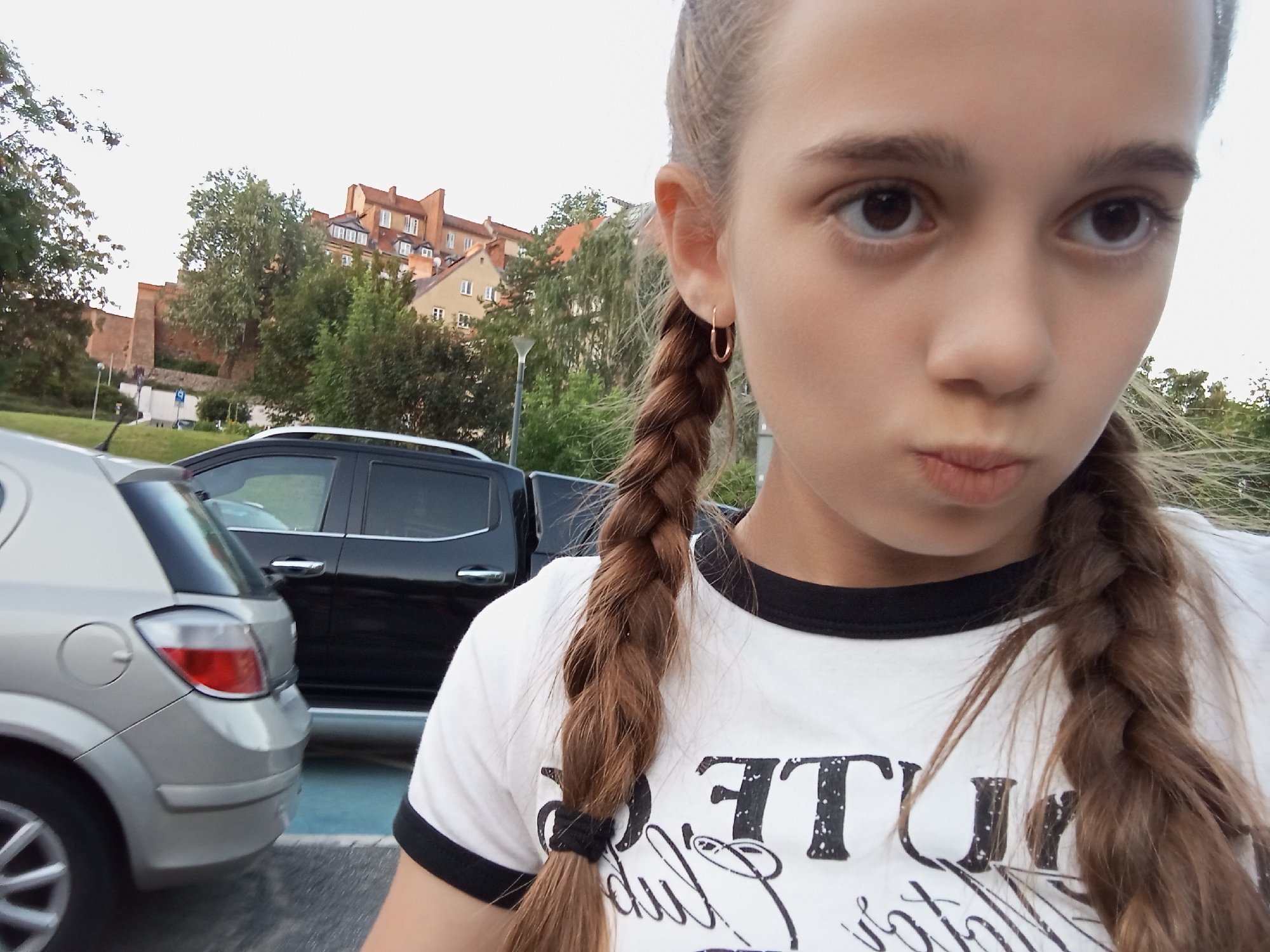 The image shows a close-up of a young girl with braided brown hair, wearing a white t-shirt with black trim and a graphic print. She is looking directly at the camera with her lips pursed. Behind her, two cars are visible: a silver hatchback on the left and a black SUV on the right. In the background, there are trees and buildings, including what appears to be a red-tiled roof and brick structures, suggesting an urban setting. The architecture of the buildings and the presence of a street lamp in the background hint at a location in Warsaw, Poland, possibly an older part of the city given the style of the buildings. The overall lighting suggests it is daytime, likely late afternoon or early evening.