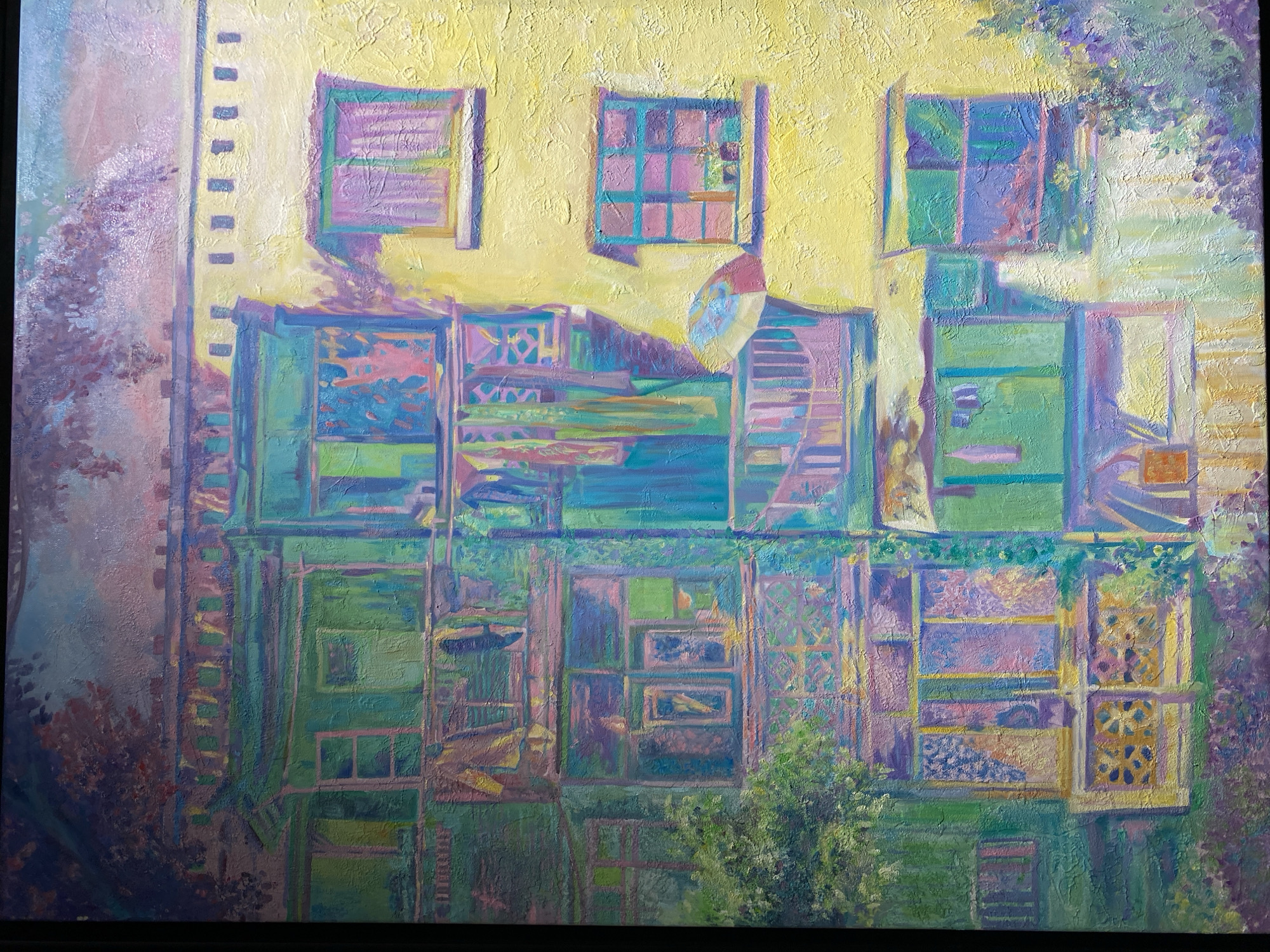 The painting depicts a vibrant, textured portrayal of an apartment building facade, heavily influenced by the distinctive architecture and atmosphere of Yuzhong District in China. The building's exterior is a riot of color, primarily featuring a bright yellow wall that contrasts with various shades of green, blue, and purple adorning balconies and window frames. Laundry hangs from several balconies, suggesting a lived-in, domestic scene. Sunlight casts strong shadows, creating a dynamic play of light and color across the structure. Lush greenery, including trees and flowering bushes, spills from the ground and climbs parts of the building, adding to the picturesque quality. The overall impression is one of urban life rendered with an Impressionistic, almost dreamlike, sensibility.