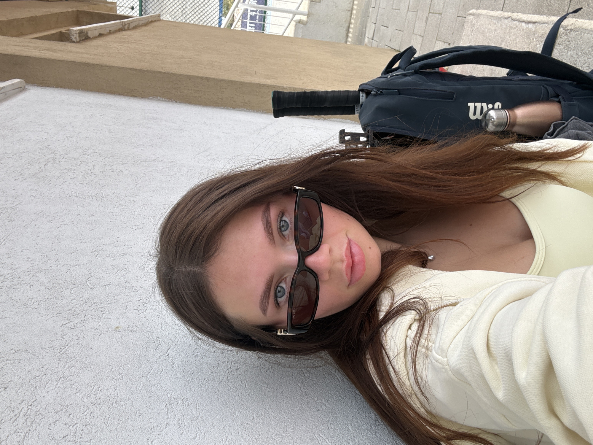 A young woman with blue eyes and long brown hair wearing sunglasses lies on a textured white surface. She is dressed in a light yellow top and an off-white jacket. A black tennis racket bag with the Wilson logo is visible in the background. The location is likely a terrace or rooftop, with a building and fence visible in the background. This image cannot be geolocated to Tel-Aviv specifically.