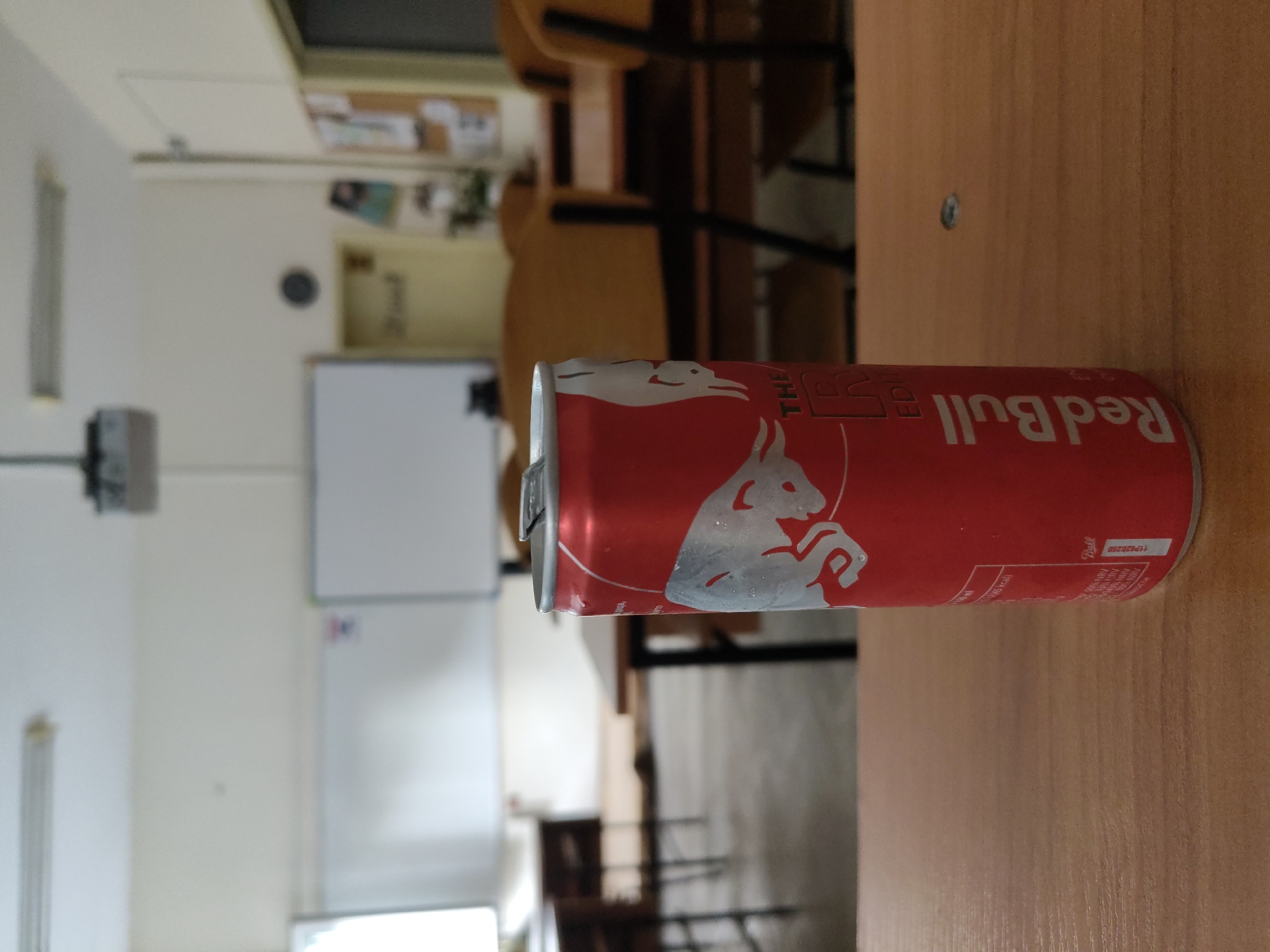 A red can of Red Bull energy drink is positioned on a wooden desk in what appears to be a classroom or lecture hall. The can is condensation-covered, suggesting it is cold. In the background, a projector is mounted on the ceiling, and whiteboards are visible on the walls. Empty chairs and tables are arranged in rows, indicating the space is unoccupied. The lighting suggests it is daytime. The image does not contain information to identify the location as Plovdiv, Bulgaria.