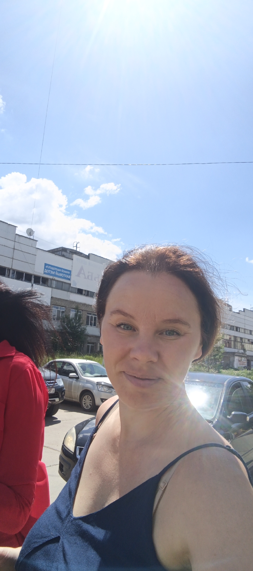 This is a medium shot, taken outdoors on a sunny day, possibly in the afternoon, with a clear blue sky and scattered clouds. The foreground is dominated by the upper body of a woman with brown hair, wearing a dark blue sleeveless top. She is smiling and looking towards the camera, with a slight lens flare reflecting on her skin. To her left, a portion of another person wearing a bright red top is visible, with their dark hair obscuring much of their face.
In the background, there is a long, multi-story building with a light-colored facade, featuring numerous windows and some visible signage. One sign appears to be in Cyrillic script and reads "ДОГЛИН НІВОТХАЯ". Several cars are parked in front of the building, including a white sedan and a dark-colored SUV. The presence of cars and the building suggests an urban or semi-urban setting. The overall impression is that of a casual, everyday moment captured on a bright, clear day. The location context is Chaikovsky, Russia.