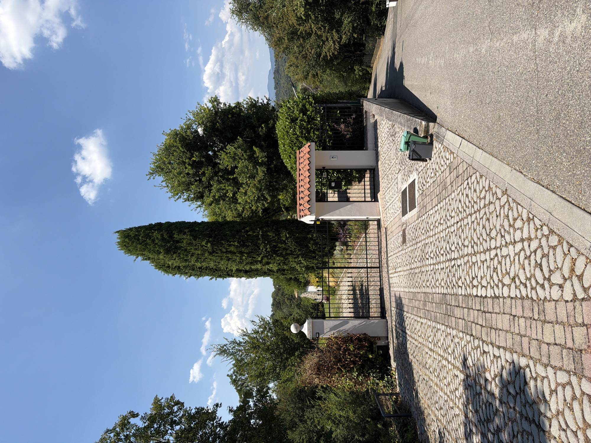 This is a vertical, eye-level, outdoor photograph of a driveway leading to a gated entrance. The driveway is paved with cobblestones and has a central strip of red brick pavers. A tall, slender cypress tree stands to the right of the driveway, and other mature trees and lush green bushes surround the property. The sky is blue with scattered white clouds. To the right of the driveway, there is a wall and a paved road. Two trash bins, one green and one black, are placed on the edge of the road. In the distance, a house with a white roof and a red-tiled entryway can be seen through the gate. The landscape beyond the property appears hilly and wooded. The scene is brightly lit by the sun, casting shadows on the driveway. The location is identified as Padenghe sul Garda, Italy.