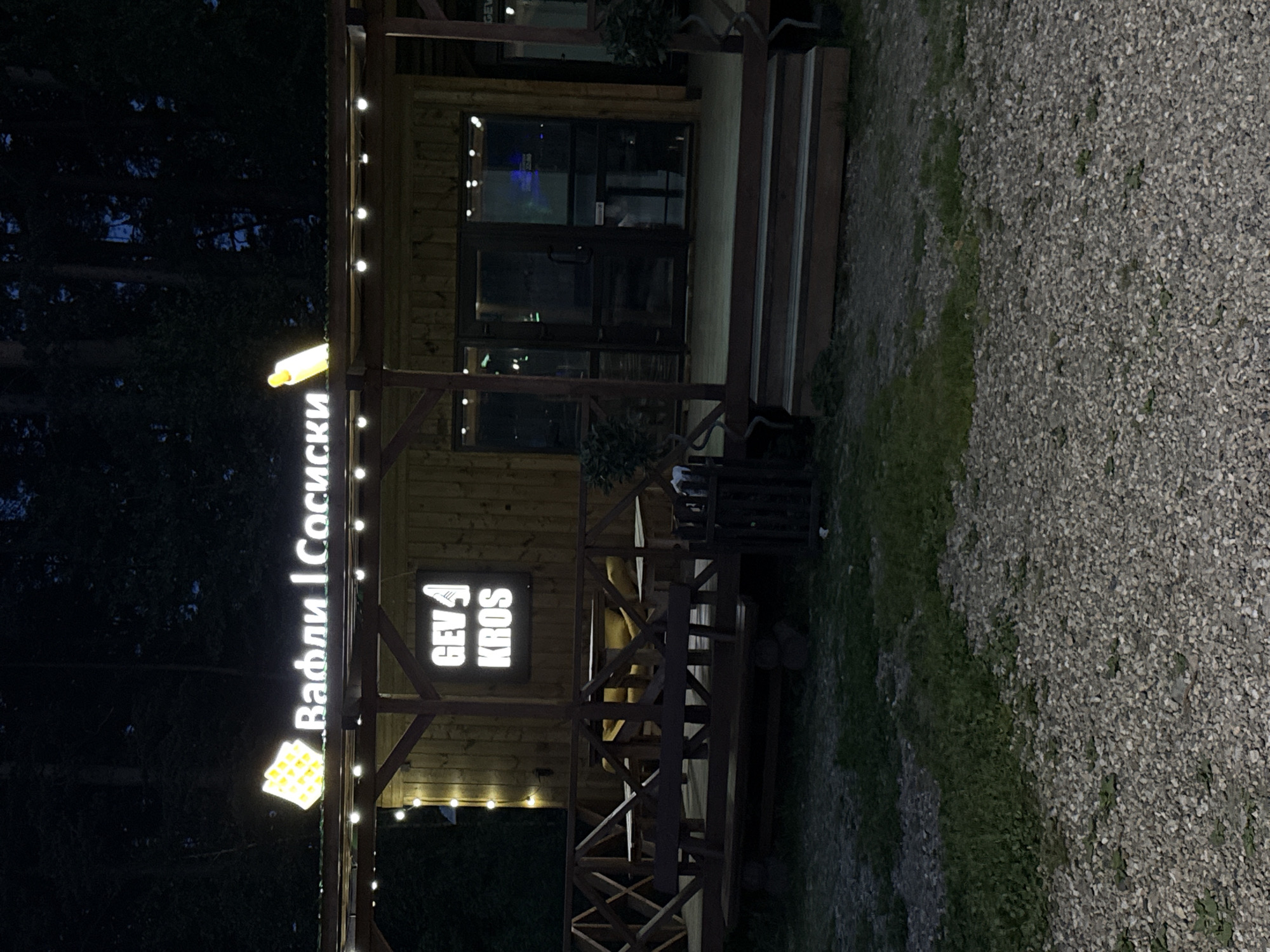 The image shows a wooden building with illuminated signs. The building appears to be a food establishment. The sign on the left reads "Вафли | Сосиски" in Cyrillic, which translates to "Waffles | Sausages." A waffle icon is positioned below the sign. Another sign on the building reads "GEV 4 KROS." The building is adorned with string lights. To the left, dense foliage and trees suggest a wooded area. To the right of the building is a gravel area with patches of grass. The location is likely Dom Otdykha Zolotoy Plyazh, Russia, given the signage in Cyrillic.