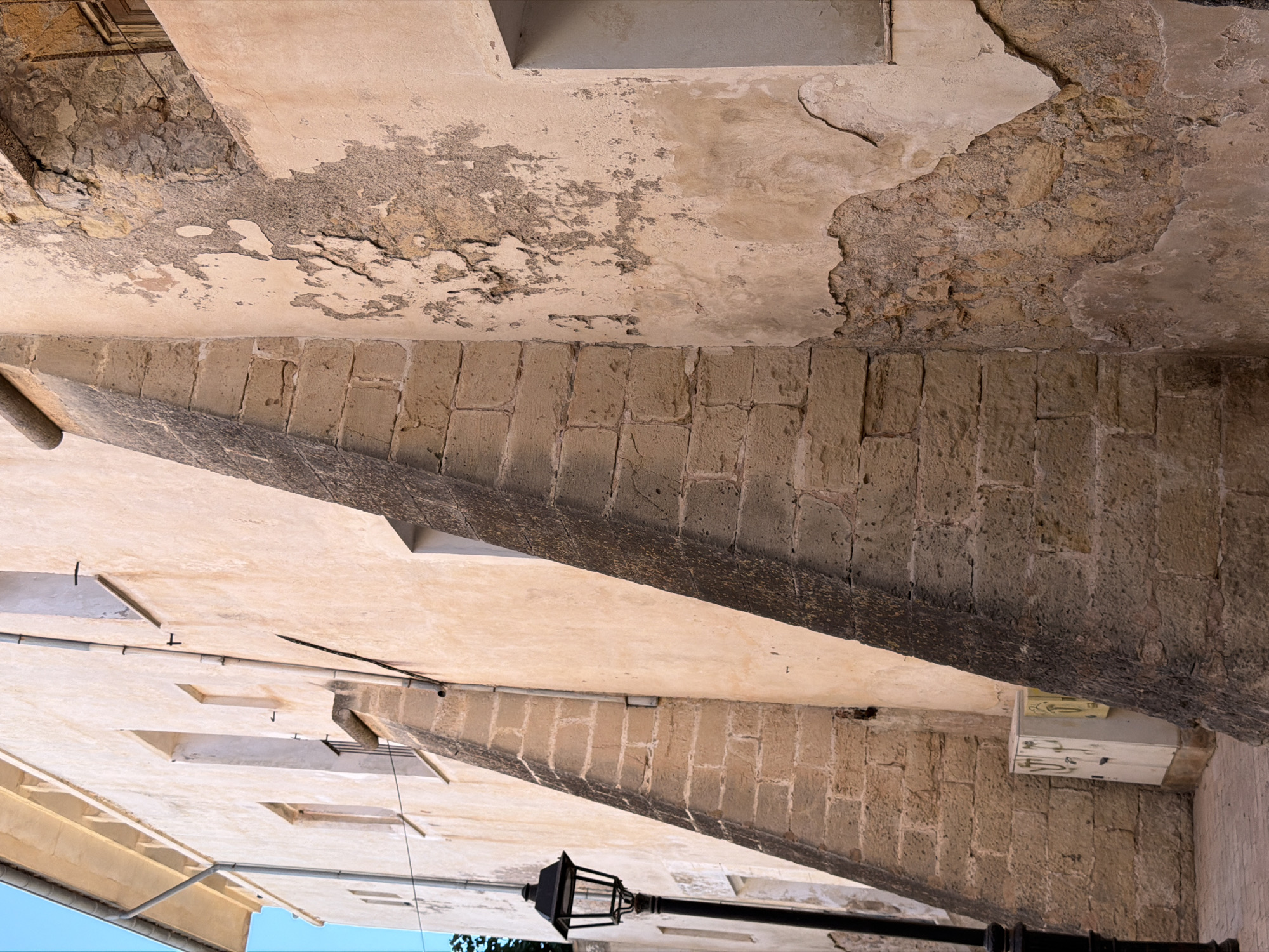 The image depicts an exterior view of a weathered building, possibly an old church or historical structure, located in the city of Manacor, Spain. The building's facade displays a combination of rough-hewn stone blocks and plaster that is peeling and deteriorated in several areas, revealing older stonework beneath. The architecture features sharp angles and buttress-like structures, suggesting an older construction style.
On the left side of the image, a black street lamp with a lantern-style fixture stands prominently. Behind it, a light-colored wall with several windows is visible. A drainpipe runs vertically down the facade. Further to the left, a glimpse of a clear blue sky can be seen, along with the edge of a gutter system at the top of the building.
In the foreground, on the right, a section of the building shows significant plaster erosion, creating an abstract pattern of exposed stone. A window with a white frame is partially visible on the far right.
At the base of the structure on the left, a white electrical box with graffiti is positioned against the stone wall. The ground in front of the building appears to be paved with reddish-brown cobblestones. The overall impression is one of age and the effects of time on the urban environment.