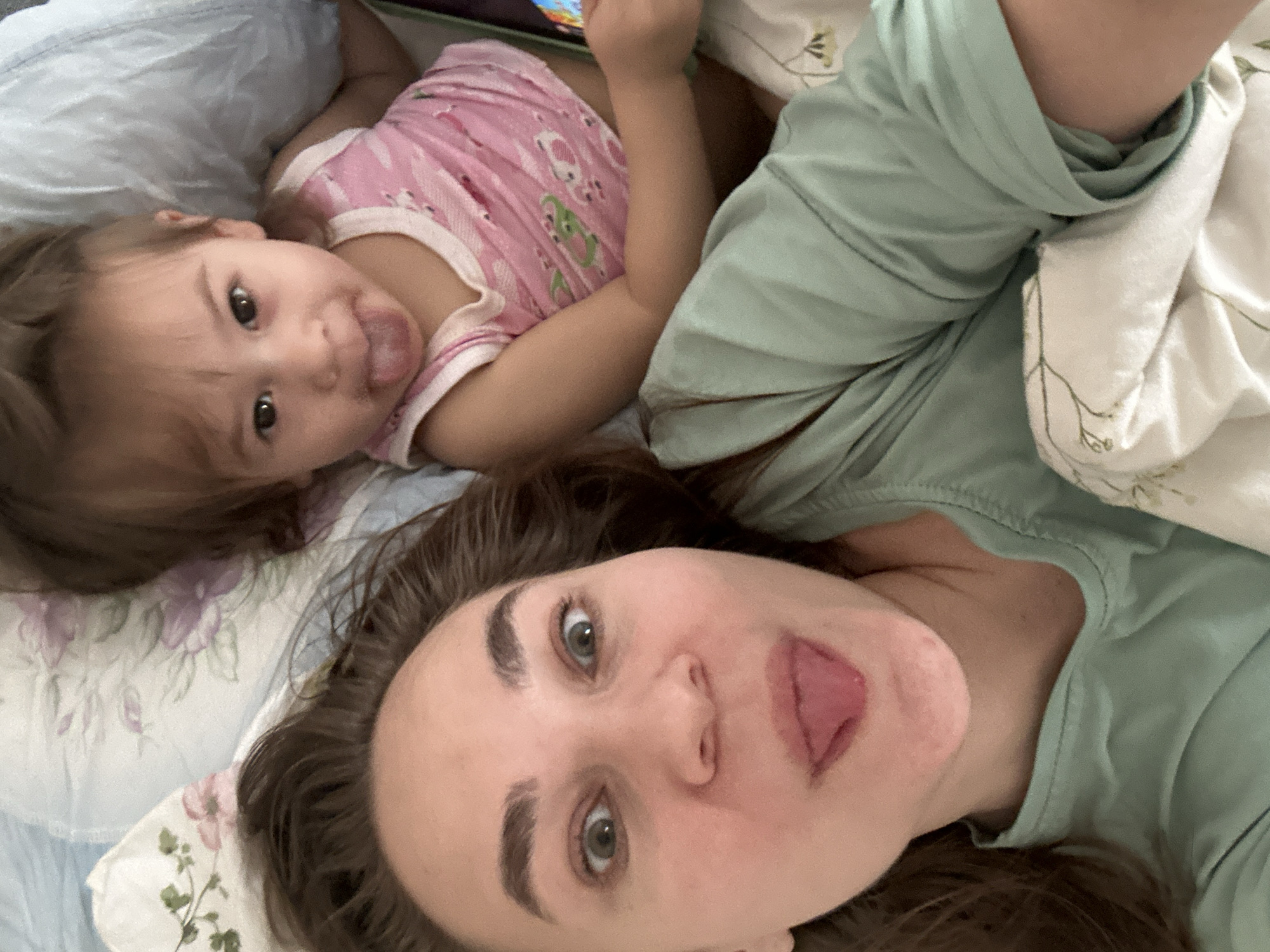 An adult and a young child are captured in a playful, close-up selfie, lying down side-by-side on what appears to be a bed. Both subjects are looking towards the camera and sticking out their tongues, indicating a lighthearted and fun moment.
The adult, positioned in the foreground and to the right, has medium-brown hair, light eyes, and prominent eyebrows. She is wearing a light green or mint-colored t-shirt. The child, situated behind the adult and to the left, has dark hair and is wearing a pink top with a pattern that includes green frog-like figures. The child's left hand is visible, seemingly holding or interacting with a screen, possibly a tablet or phone, though only a small part of it is discernible.
The setting appears to be indoors, likely a bedroom, with soft bedding visible. The adult is partially on a white pillow with subtle green foliage patterns, while the child is on a pillow with light blue and purple floral designs. The overall atmosphere is relaxed and intimate. The image provides no specific time of day or weather cues beyond indoor lighting. The location context for this image is Kharnavy, Russia. No visible text is present in the image.