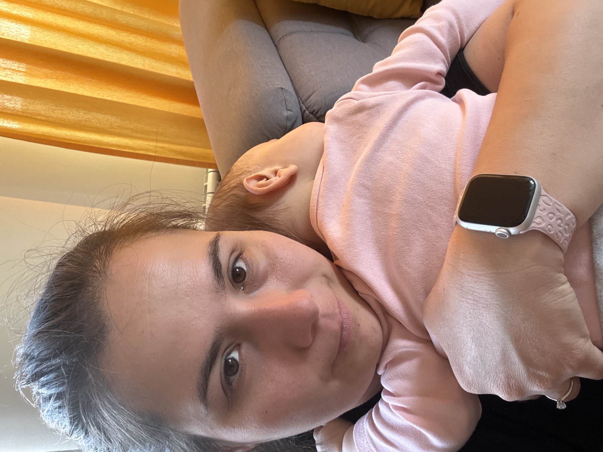 This intimate portrait, taken indoors in Sofia, Bulgaria, captures a tender moment between a woman and a baby. The image is oriented sideways, showing the subjects horizontally.
The woman, likely the mother, is in the foreground, looking towards the camera with a soft, gentle smile. She has dark hair and brown eyes, and is wearing a light pink long-sleeved top. On her left wrist, she wears a silver-colored smartwatch with a light pink, textured band, and a silver ring with a single clear stone is visible on her left ring finger.
Nestled against her chest or shoulder, the baby is largely visible from the back of the head, showing a small ear. The baby is also dressed in a light pink long-sleeved garment, seemingly asleep or resting comfortably.
The background reveals elements of a home interior. A gray upholstered couch or armchair provides a soft resting place for the subjects. Above them, a warm yellow-orange horizontal-slatted window blind suggests natural daylight illuminating the scene. The overall mood is one of peace, affection, and contentment between mother and child during the daytime. No text is visible within the image.