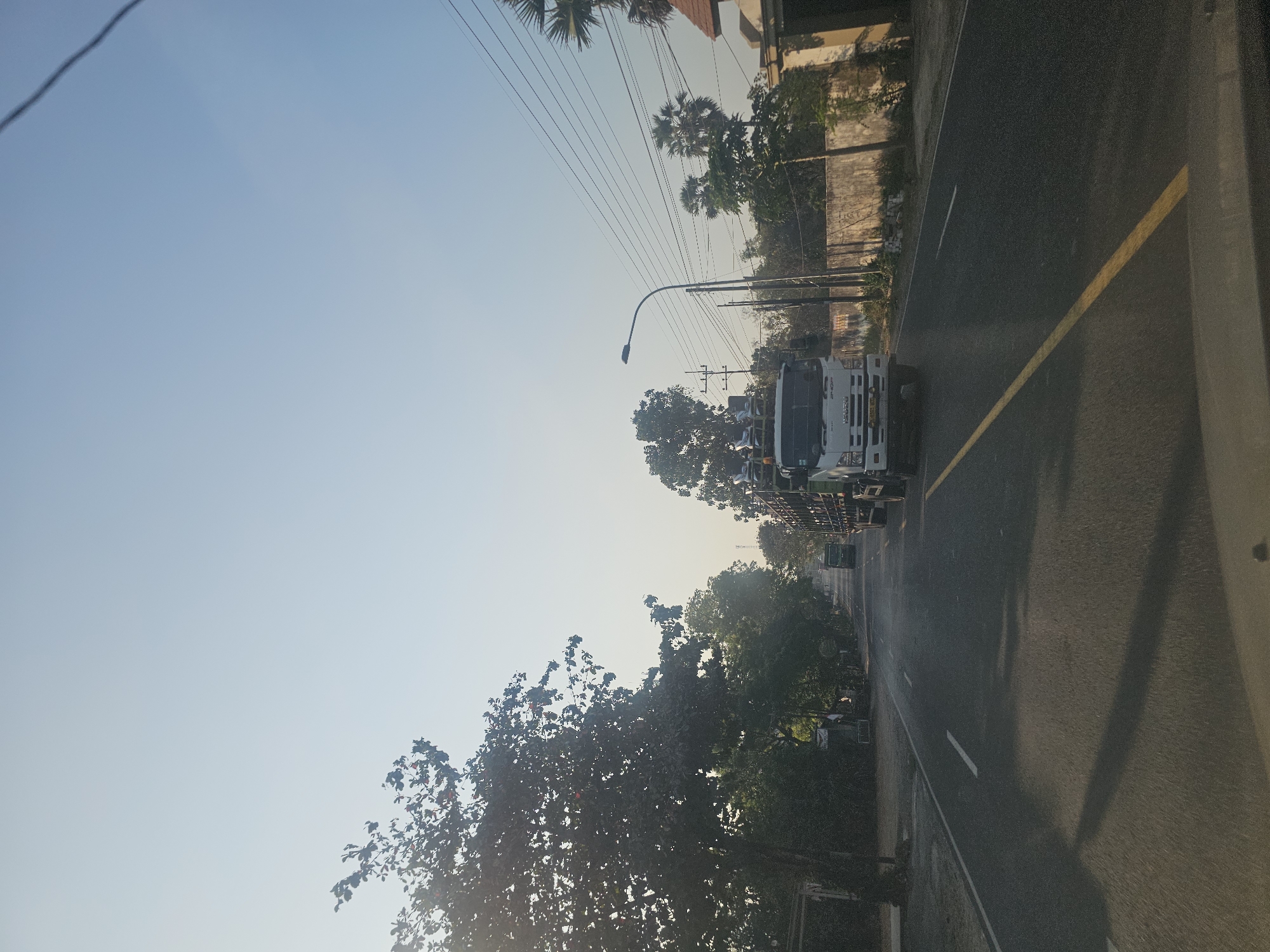 The image depicts a road in Palang, Indonesia, likely captured during the early morning or late afternoon, judging by the low angle of the sun and the soft lighting. A white truck, identified by the "ISUZU" emblem on its grille, is prominently featured driving on the right side of the road. The truck appears to be carrying cargo, possibly construction materials or produce, secured on its flatbed. In the distance, another vehicle, possibly a smaller truck or van, is visible further down the road.
The road itself is asphalt with a distinct yellow line dividing the lanes. The environment is characterized by lush greenery, with tall trees lining the sides of the road. The foliage is dense, suggesting a rural or semi-rural setting. Power lines are strung across the scene, supported by utility poles. On the right side of the road, a building with a tiled roof is partially visible, along with a concrete wall and some vegetation. The sky is a clear, pale blue, indicating fair weather. No individuals are clearly identifiable in the frame. The overall impression is one of a typical transportation scene in a tropical or subtropical locale.