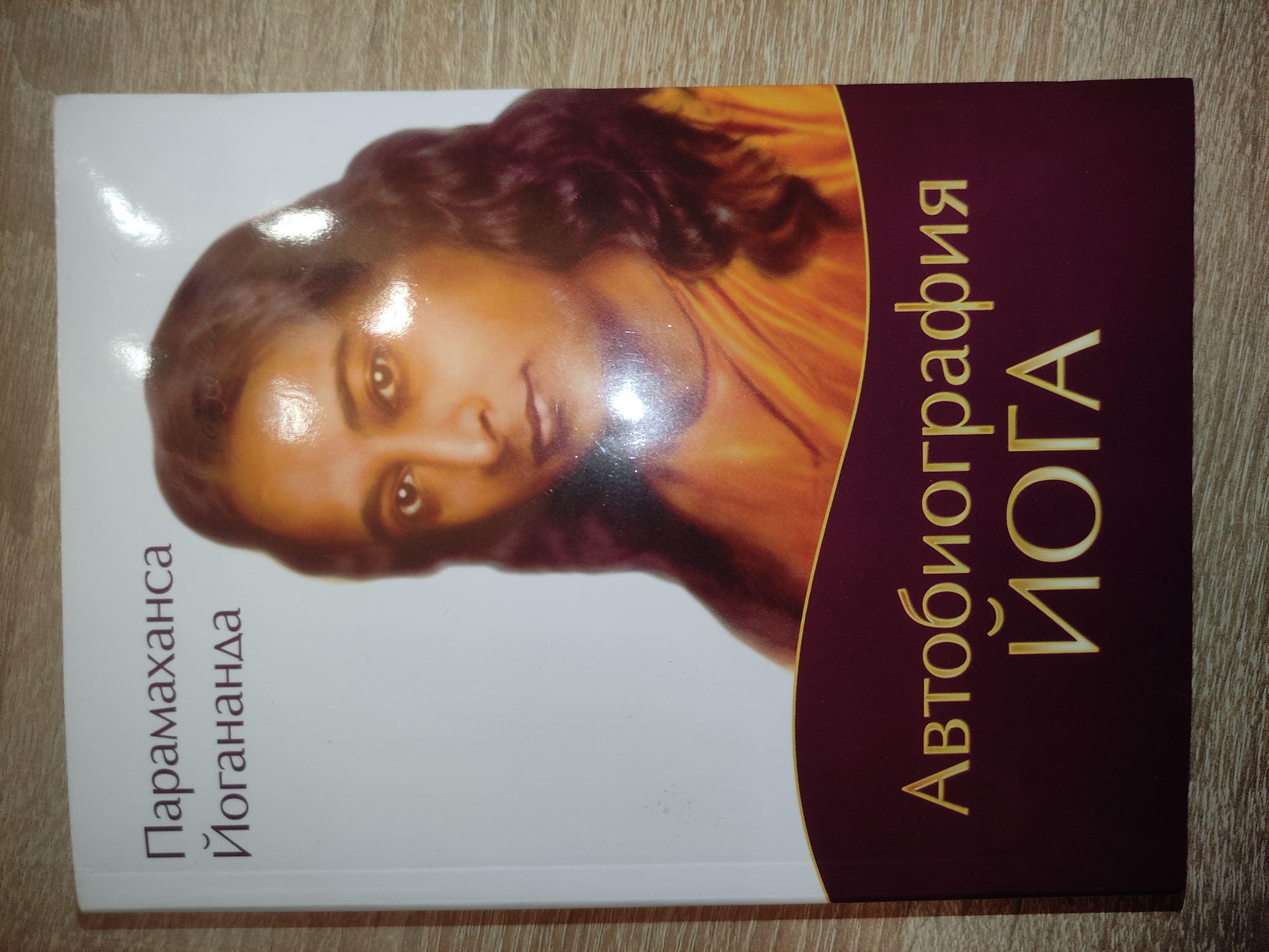 A book with a glossy cover depicting a portrait of Paramahansa Yogananda rests on a light-colored wooden surface with visible grain. The cover's upper section is white, featuring the Russian text "Парамаханса Йогананда" in brown. Below this, a portrait of a person with dark, shoulder-length hair, light brown skin, and wearing an orange garment is centered. The lower section of the cover is dark maroon, separated from the white by a thin gold curved line, and displays the Russian text "Автобиография ЙОГА" in gold, translating to "Autobiography of a YOGI." Direct light causes glare on the upper right portion of the book cover. The scene is situated in the city of Kratovo, Russia.