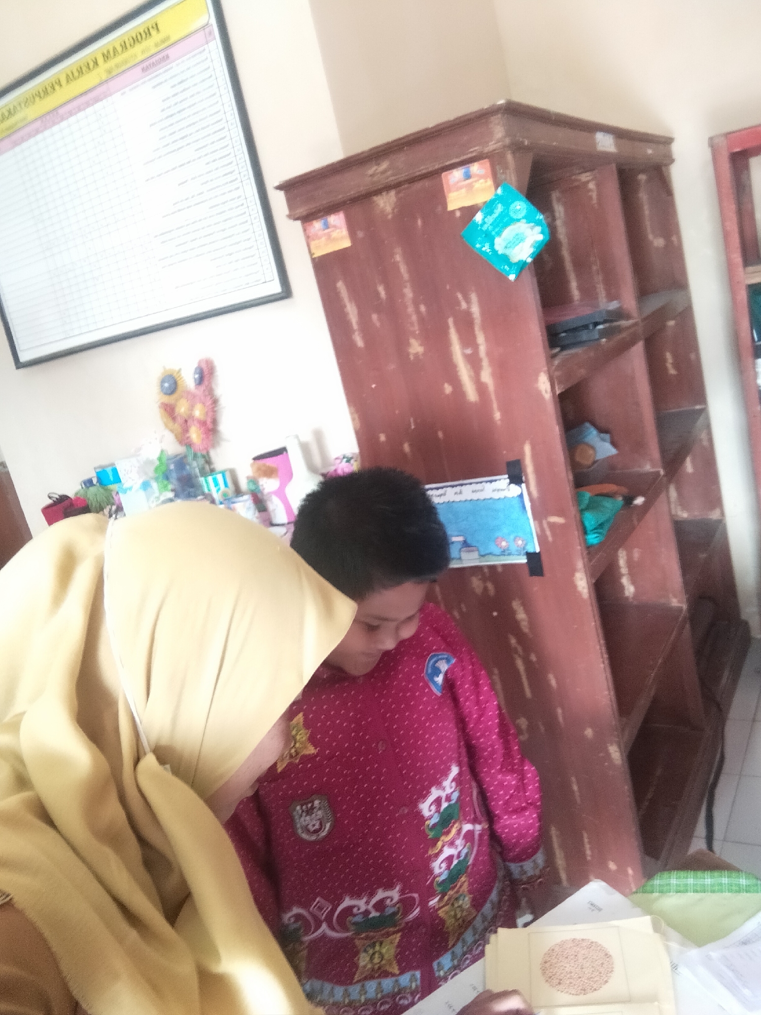 In an indoor setting, possibly a classroom or a common area in Tretes, Indonesia, a young child wearing a pink and red patterned shirt is engaged in an activity. The child's face is partially visible, looking down at a piece of paper. To the left of the child, a person wearing a yellow hijab is also looking in the same direction. The background features a large, worn wooden cabinet with multiple shelves. One shelf displays a blue and white drawing. To the left of the cabinet, a large framed chart with text and a grid is affixed to the wall. Various small decorative items and containers are scattered on a surface in front of the chart. The overall lighting suggests an indoor environment.