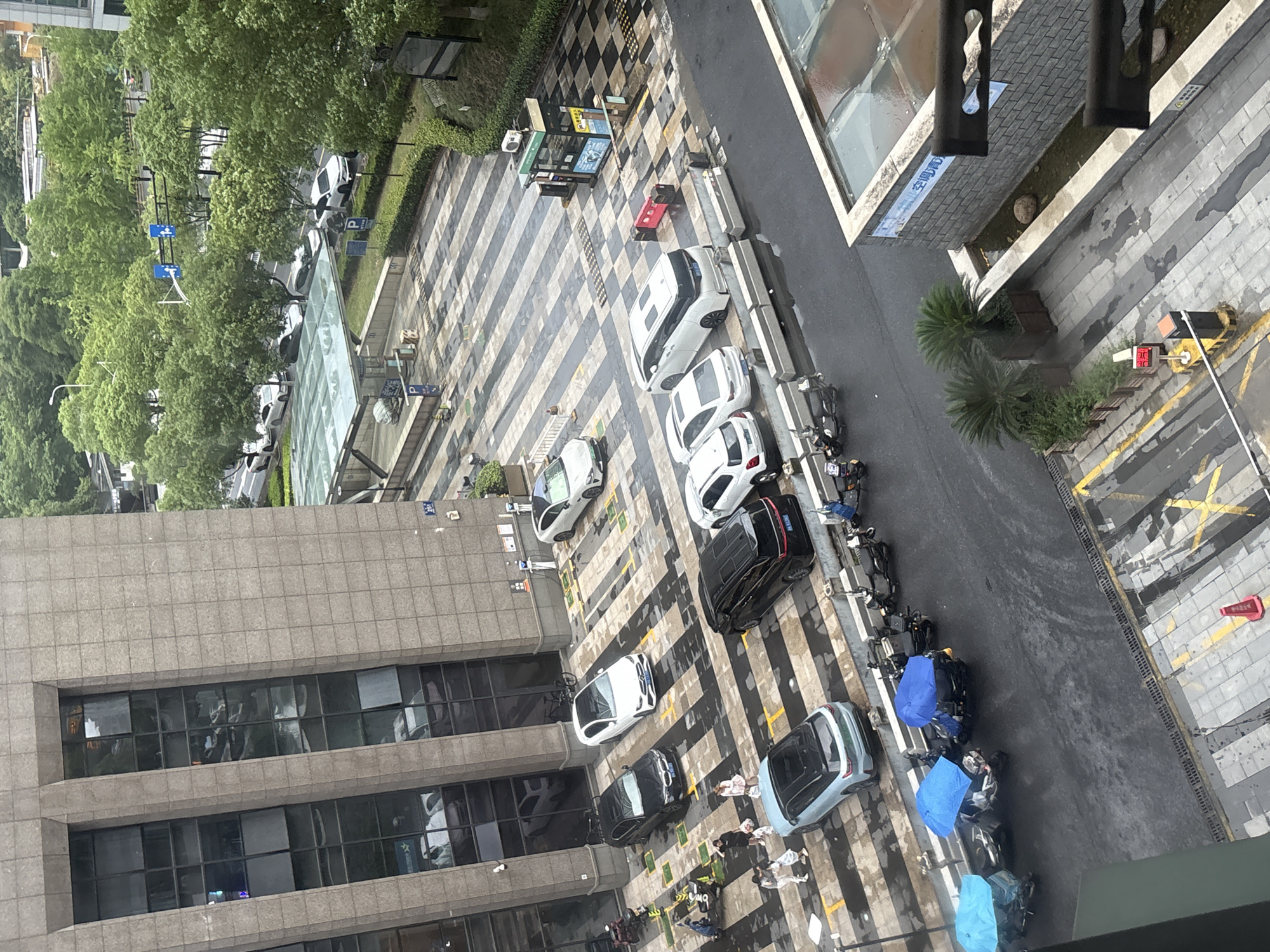 The image shows an outdoor scene in an urban environment, likely a parking area or street adjacent to a building. There are several cars and motorcycles parked. Some of the motorcycles are covered with blue umbrellas, suggesting it is raining or has recently rained, as the ground appears wet and reflective. A group of people are standing near some of the cars, and one person is walking. The building in the background has a facade made of stone tiles and features large windows. In the distance, there are trees and other buildings. There are also traffic signals and street signs visible. The overall impression is of a typical day in a city, with some individuals engaged in everyday activities and vehicles present.
