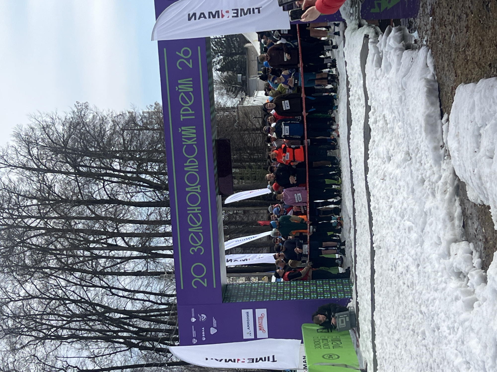 The image shows the starting line of the "20 Zelenodol'skiy Trail 26" race in Zelenodol'sk, Russia. Runners are assembled at the starting line beneath an arch displaying the race name in Russian and the word "TIME:MAN". There is a large snowbank to the right of the starting line. Bare trees are visible in the background, as is a building. Several flags with the word "TIME:MAN" written on them are displayed on the left. An individual is crouched behind a small green table in the lower right corner. Spectators are observing the race.