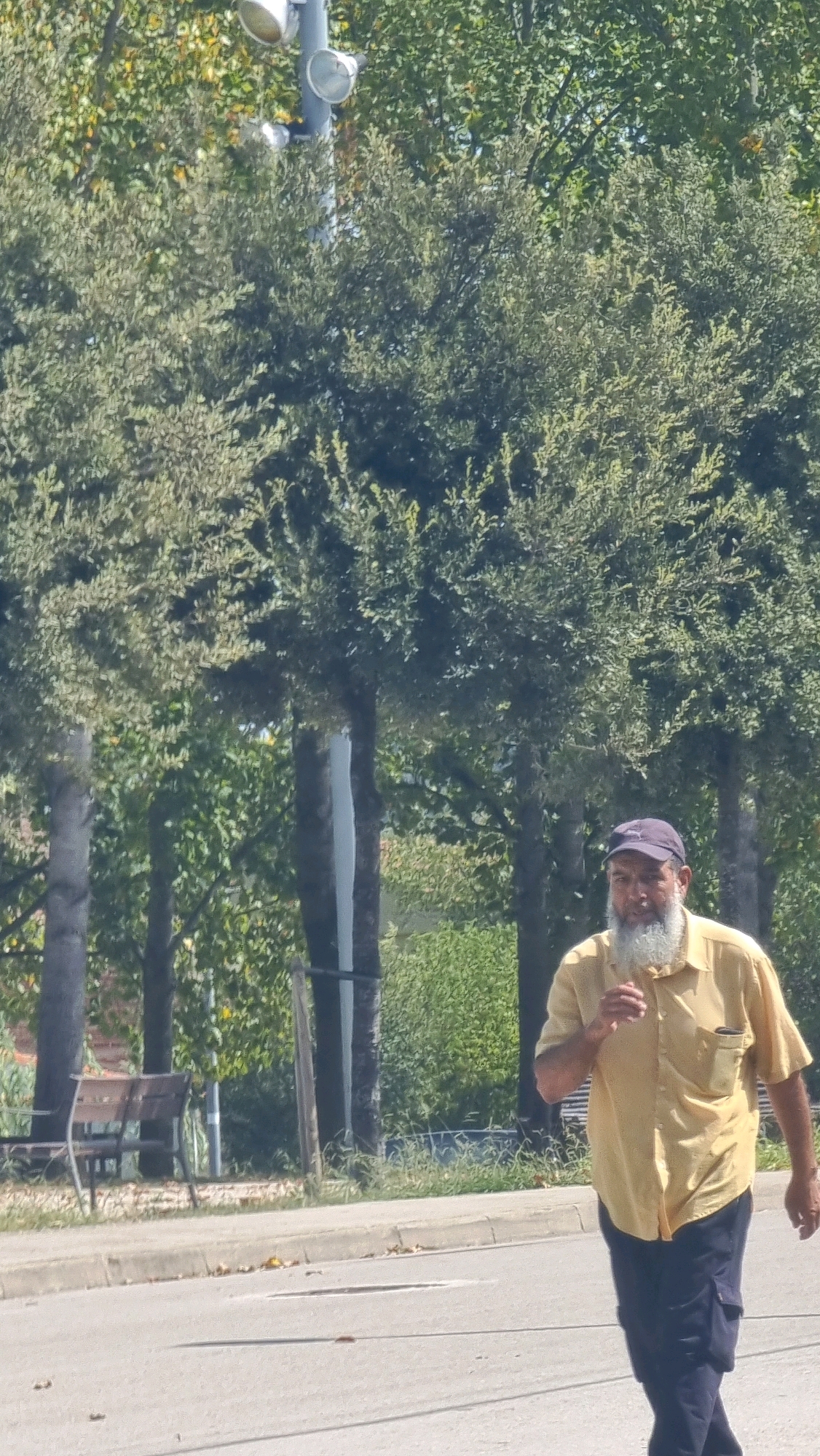 An adult male with a full white beard and wearing a yellow collared shirt and dark pants is walking on a paved road. He is wearing a dark baseball cap. Behind him, a sidewalk with some grass and a bench are visible. In the background, there are trees and a lamppost with two lights. The scene appears to be outdoors in a park-like setting. The location is identified as Sant Joan les Fonts, Spain.
