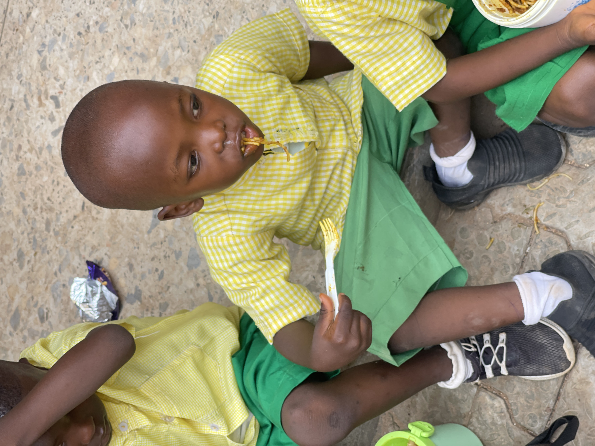 A young boy, wearing a yellow and white checkered shirt and green shorts, is eating spaghetti with a white plastic fork. He is sitting on the ground, which appears to be paved with interlocking stones. Another child, also wearing a similar uniform, is partially visible to the left of the frame. A plastic container with spaghetti and a foil-wrapped item are also on the ground. The scene appears to be outdoors, possibly in a schoolyard or common area. The lighting suggests it is daytime. The overall impression is one of a mealtime in a community setting. The location is Akwanga, Nigeria.