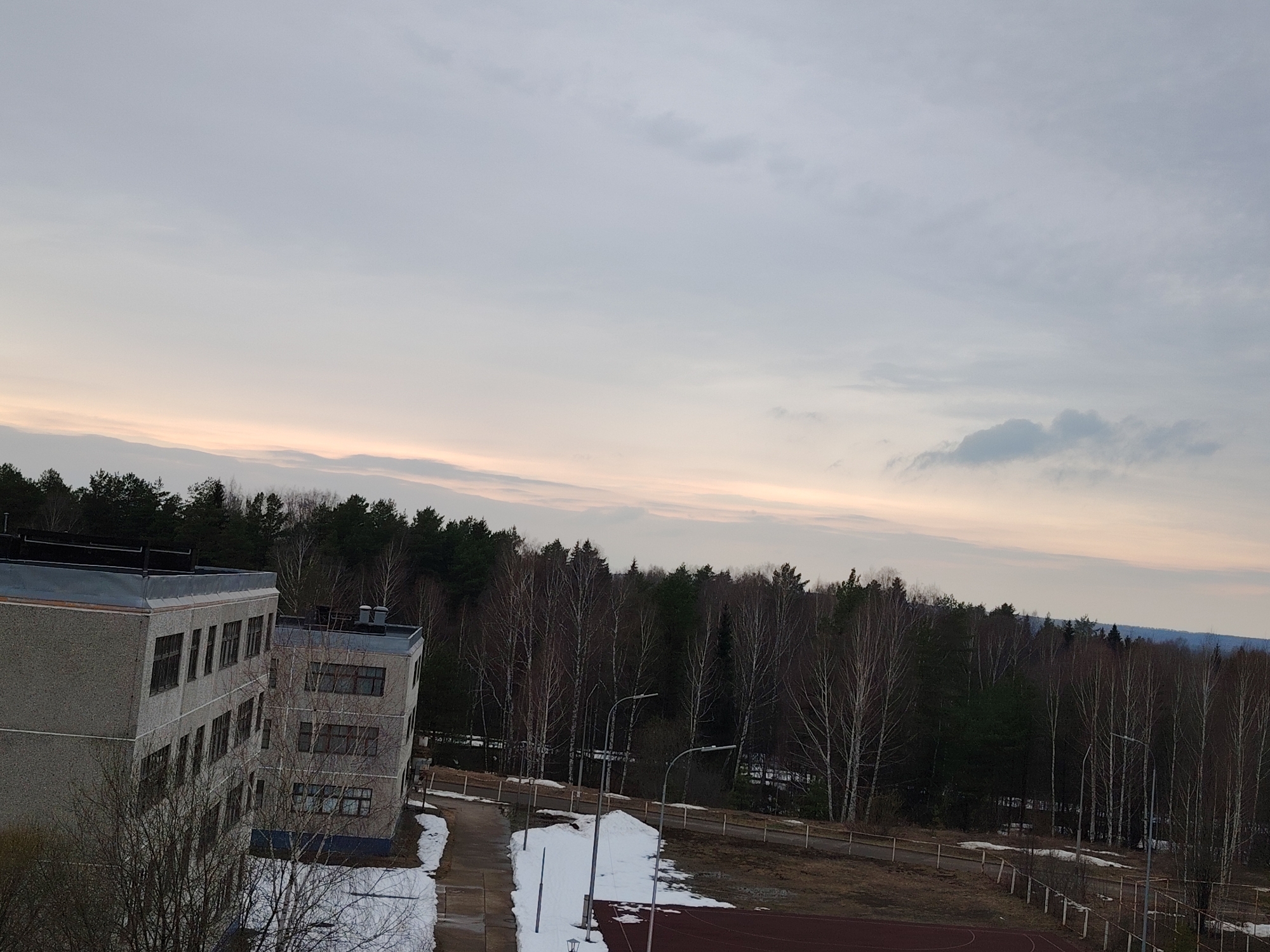 The image shows a view of a building and surrounding landscape in Марковский, Russia. The sky is overcast with hints of pink near the horizon. A large, multi-story gray building is visible on the left, featuring multiple windows. Patches of snow cover the ground around the building, and bare trees are interspersed with evergreen trees, forming a forest in the background. Street lights are positioned along a walkway near a sports field, partially visible at the bottom. A fence runs parallel to the walkway. The landscape appears to be in a transitional season, between winter and spring.