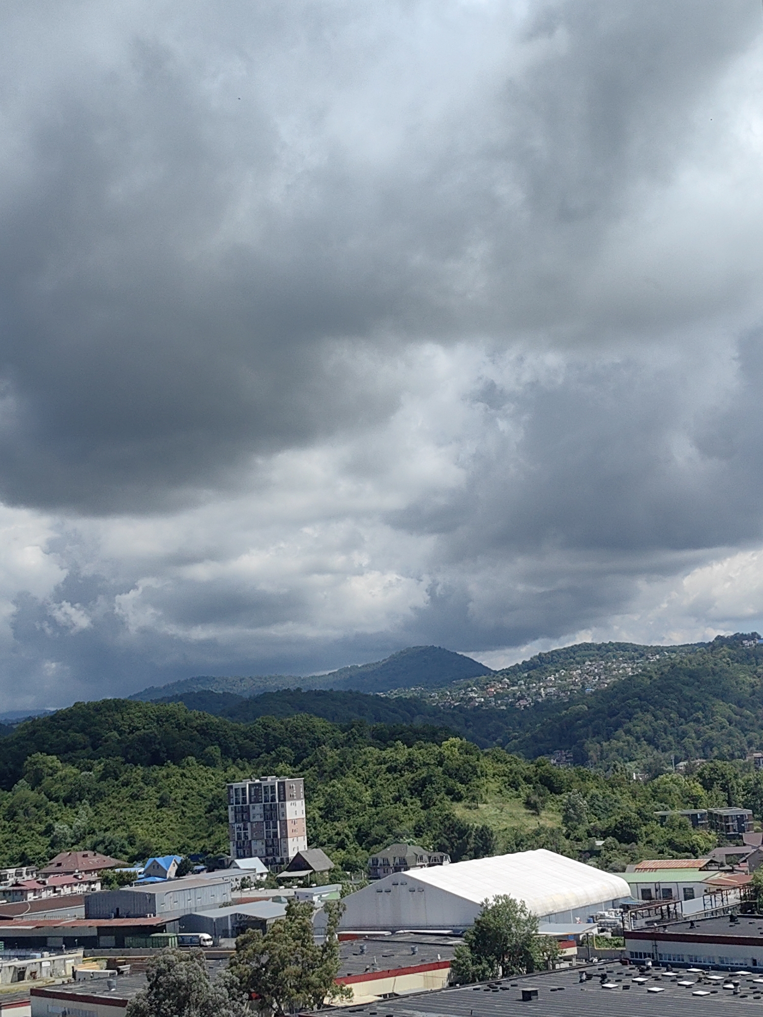 The image shows a cloudy sky over a city with buildings nestled in a valley and on hills. In the foreground, there are several low-rise buildings with flat roofs, some industrial-looking structures, and a large white tent-like building. A few trees are visible among the buildings. In the middle ground, a tall, multi-story building with a checkered facade stands out against a backdrop of lush green hills. The hills are densely covered with trees, and houses are scattered across the slopes, creating a village-like appearance. In the background, several mountain ranges are visible, receding into the distance under a thick layer of dark, dramatic clouds. The overall impression is of a settlement within a mountainous, possibly humid, environment. The scene is consistent with the terrain and architecture found in Sochi, Russia, a city known for its coastal location and proximity to the Caucasus Mountains, where such cloud formations and dense vegetation are common.