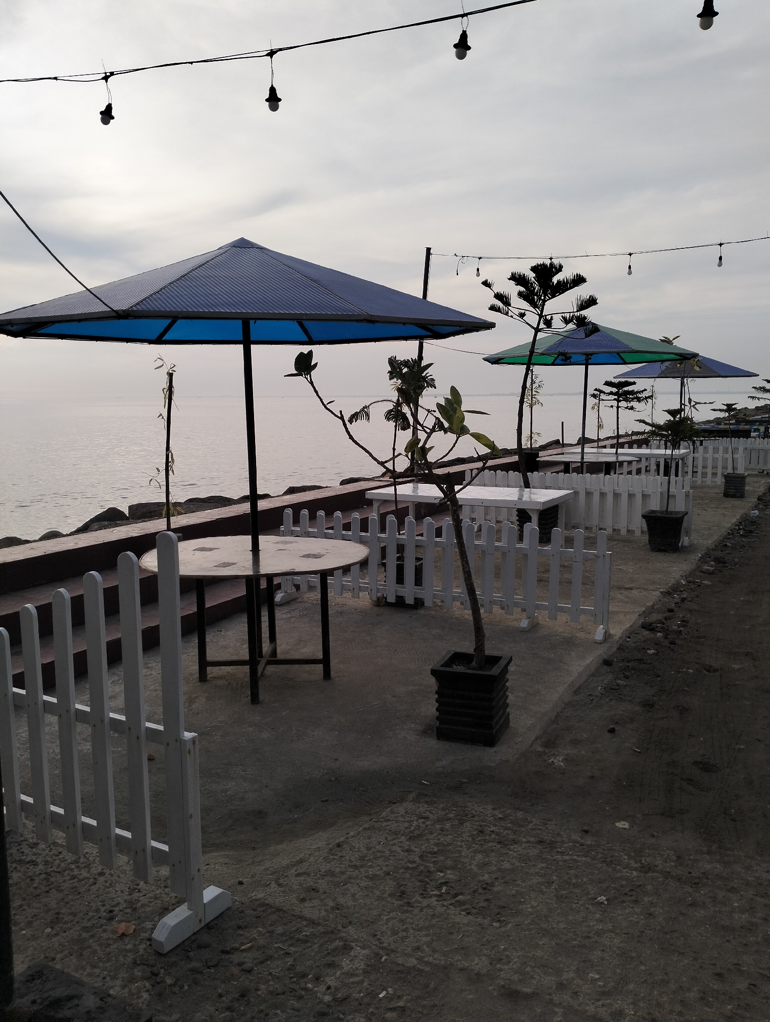 The image depicts a seaside outdoor seating area in Lhokseumawe, Indonesia. The scene includes a series of round tables and rectangular tables with black metal legs, each covered by a large, blue or blue and green umbrella. A low, white picket fence runs along the seating area, separating it from the shoreline. The seating area is lined with potted plants in black square pots. Overhead, strings of lights are suspended. The sea stretches out to the horizon under an overcast sky. The ground is a mixture of concrete and dirt. The scene appears unoccupied.
