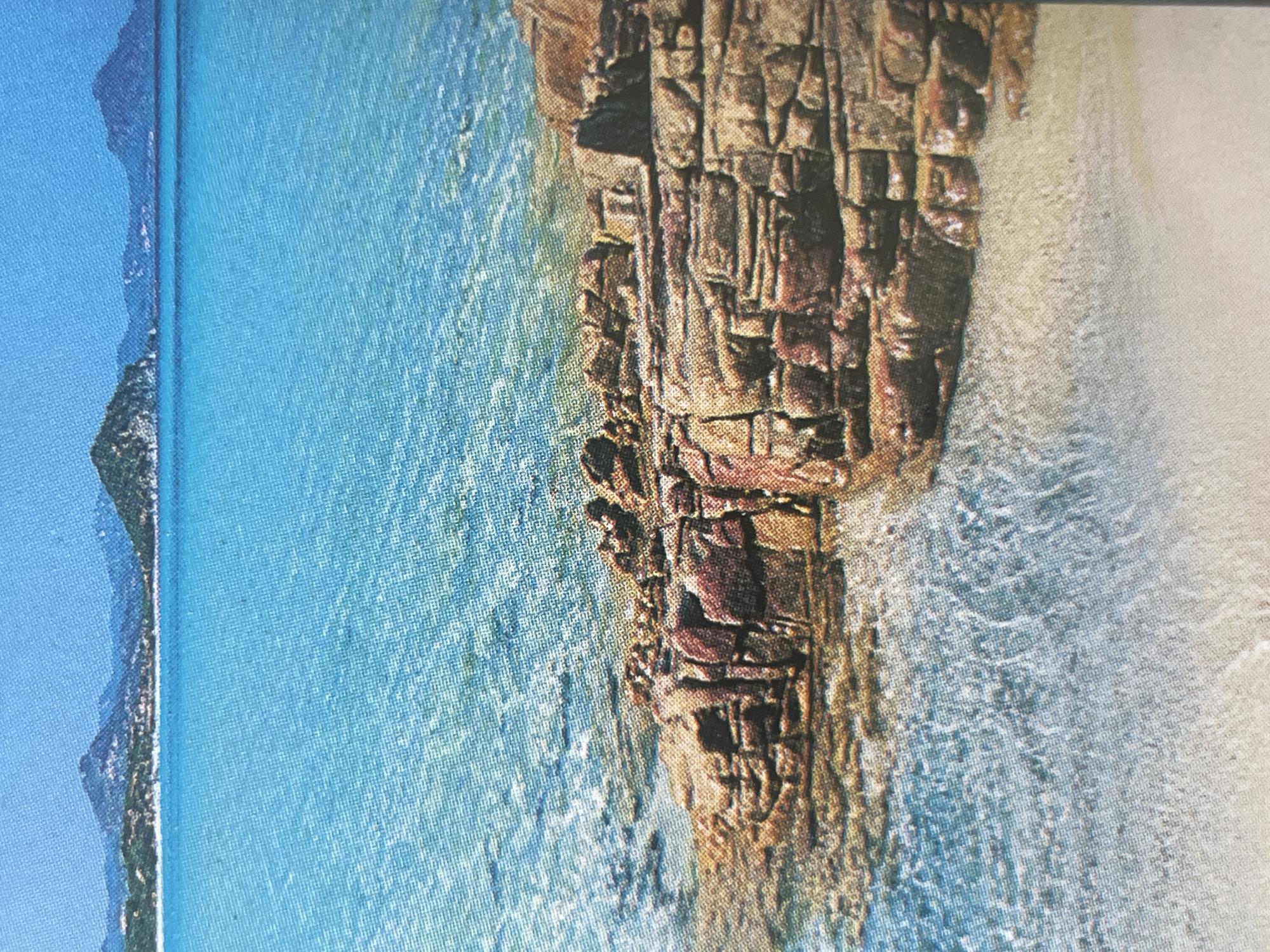 The image depicts a coastal landscape with a clear blue ocean in the foreground, meeting a sandy beach with prominent rock formations. In the distance, a coastline with mountains and scattered buildings is visible under a bright blue sky. There are no people or identifiable structures in the image. The scene is a natural, serene environment. This image is not related to the city of Sofia, Bulgaria.