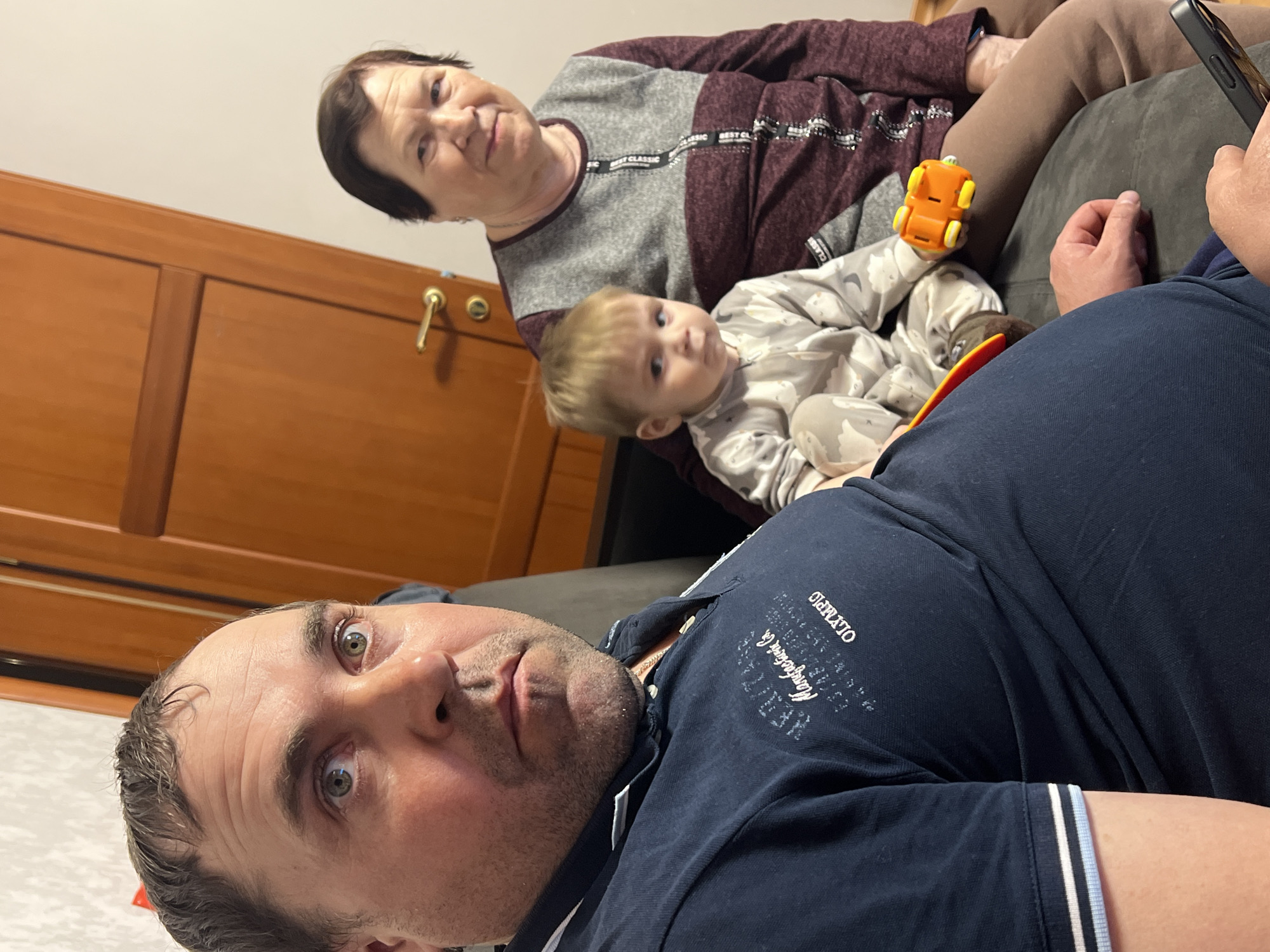 This indoor photograph, taken in Perm, Russia, captures three generations of a family in a casual setting. In the foreground, a man with short dark hair and stubble is seen from a low, tilted angle, looking directly at the camera with wide, expressive eyes. He wears a dark blue polo shirt, which features partially visible text including "OLYMPIC" and "Manufacturing Co." His arm and hand are visible on the far right of the frame, holding a black smartphone, suggesting he is either taking a selfie or being photographed.
Behind him, an older woman with short dark hair and a gentle smile is seated, looking towards the camera. She wears a gray and maroon long-sleeved top adorned with "BEST CLASSIC" text. Cradled between the woman and the man's arm is a young child with blonde hair, dressed in a light-patterned onesie. The child holds an orange toy car with yellow wheels and also looks curiously towards the camera. The family appears to be relaxing on a dark gray sofa. In the background to the left, a wooden door or piece of furniture with a gold-colored handle is visible. The scene conveys a warm, intimate family moment captured under indoor lighting.